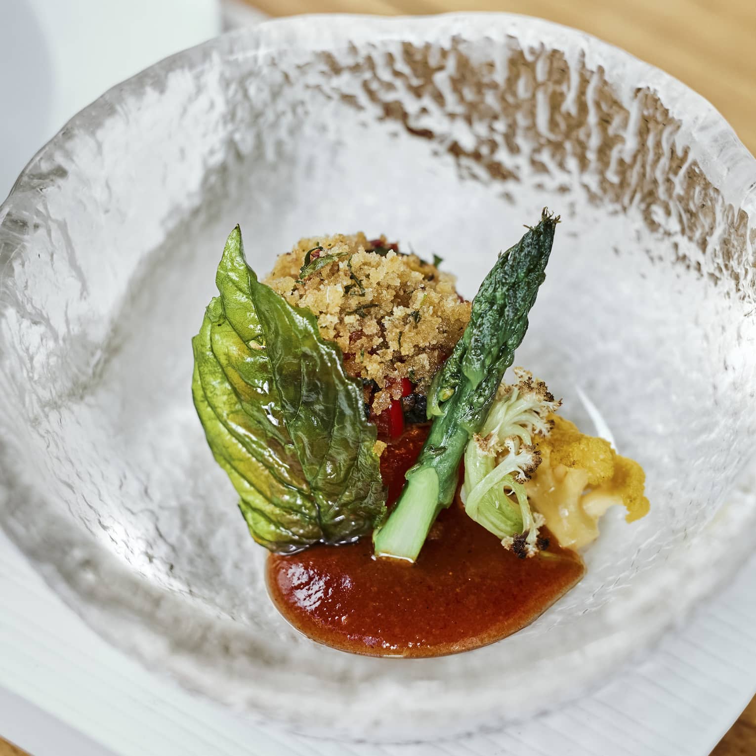 A croquette rests in a textured crystal bowl with red sauce, small grilled vegetables and a translucent fried herb. 