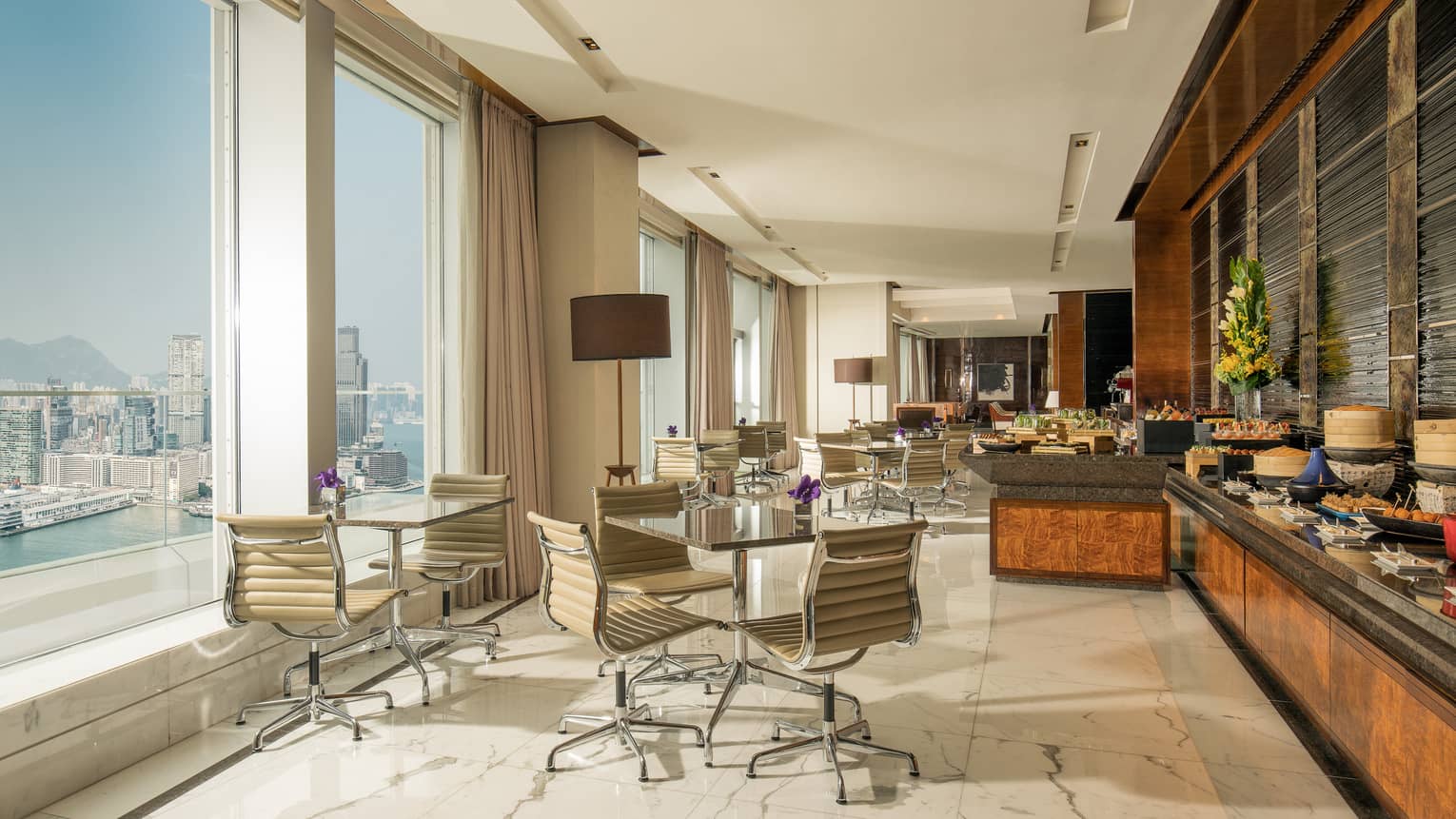 Hotel lounge with wheeled leather desk chairs at round tables across from buffet and beside tall windows with water view