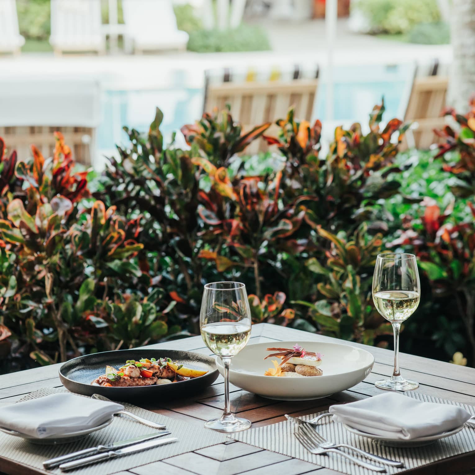 Outdoor dining table with two plated meals and two wine glasses, with foliage and a pool in the background.
