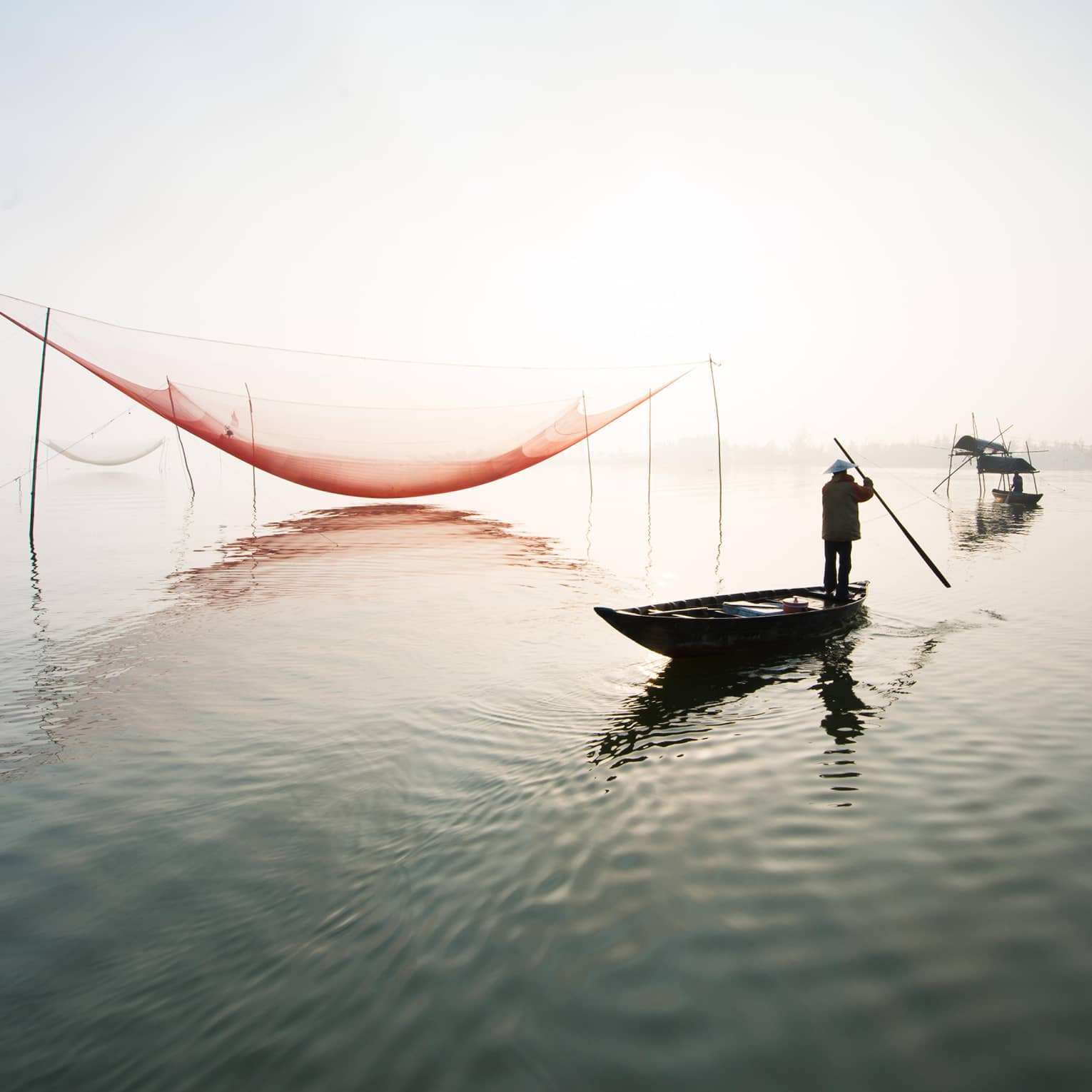 A man fishes standing up in a canoe as the sun shines brightly behind him. 