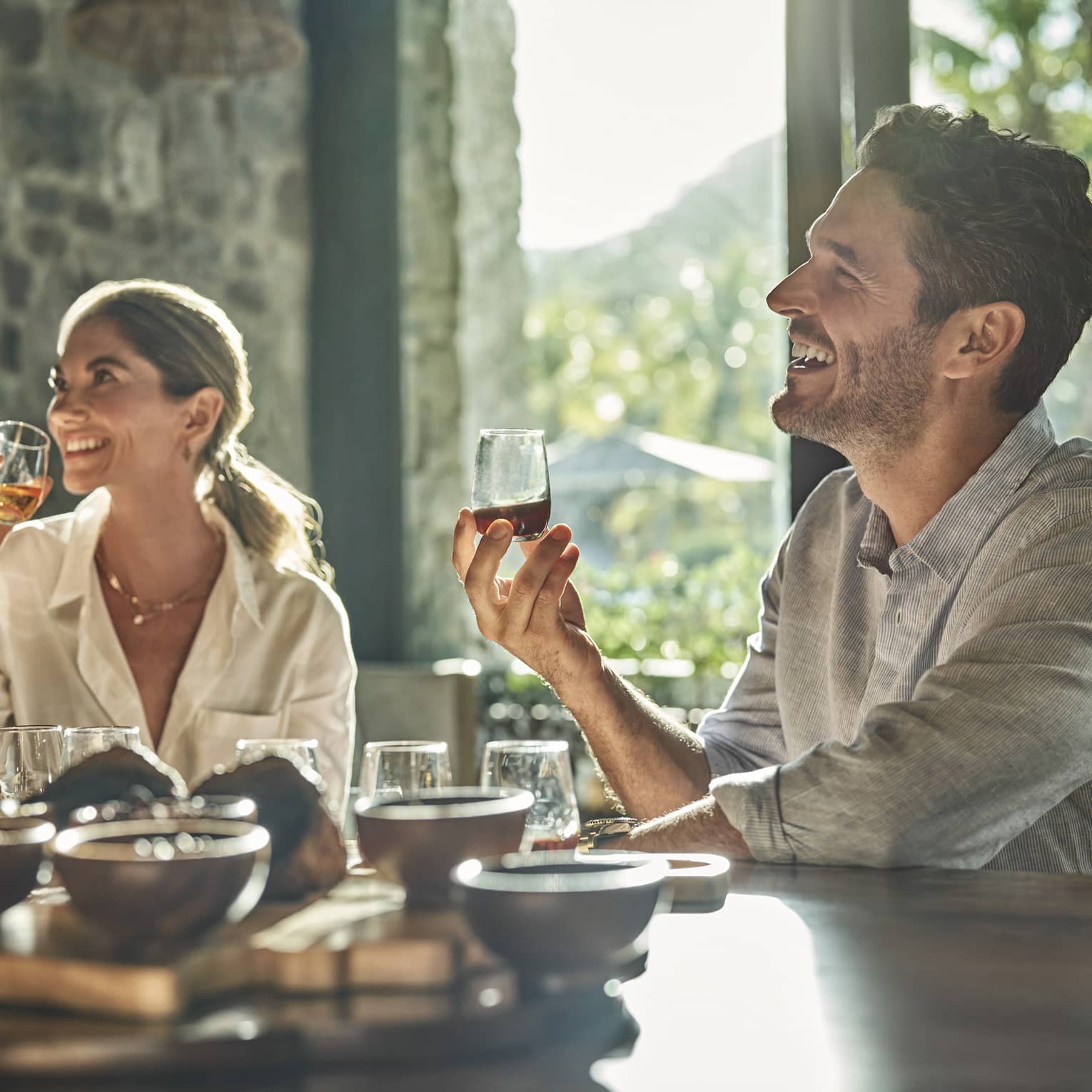A man and a woman sit at a counter looking up and smiling and each holding a glass of rum with various other glasses and ingredients sit in front of them on the counter