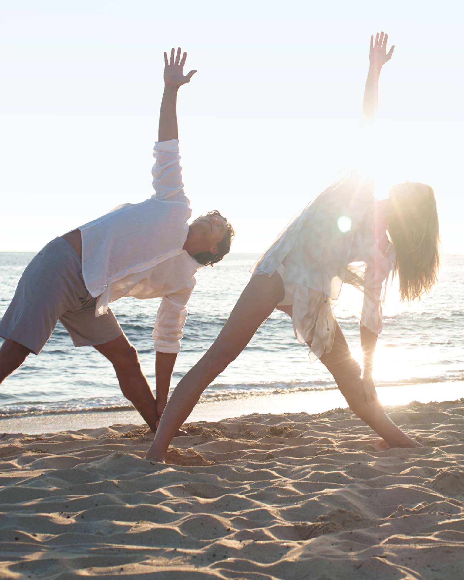 Two people in white beach shirts and shorts do a yoga pose with yogi at sunrise on beach
