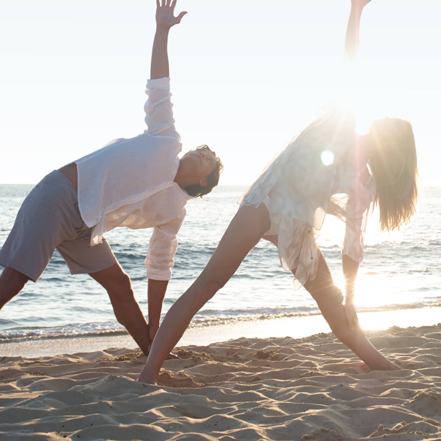 Two people in white beach shirts and shorts do a yoga pose with yogi at sunrise on beach