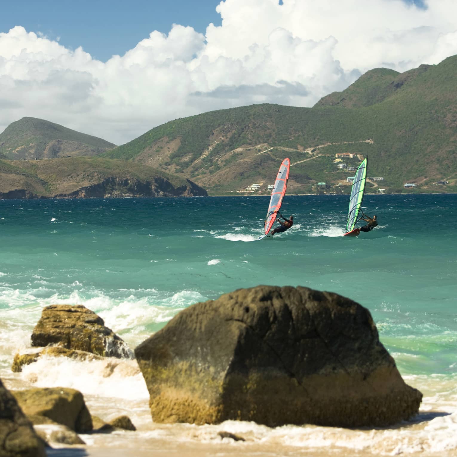 Two windsurfers glide on waves in bay between rocks, green mountains