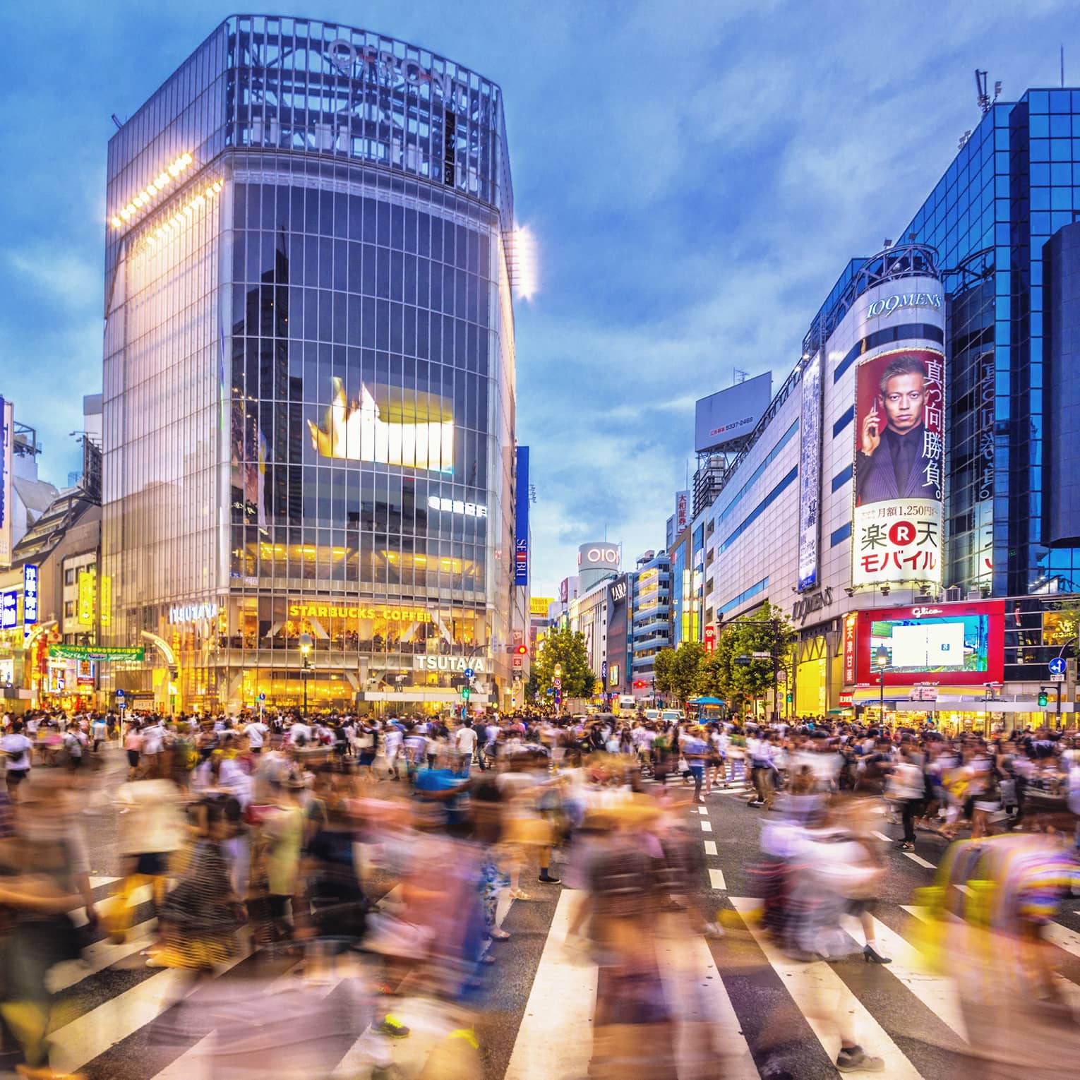 Image of blurry crowds along sidewalk under glass buildings, colourful billboards at night
