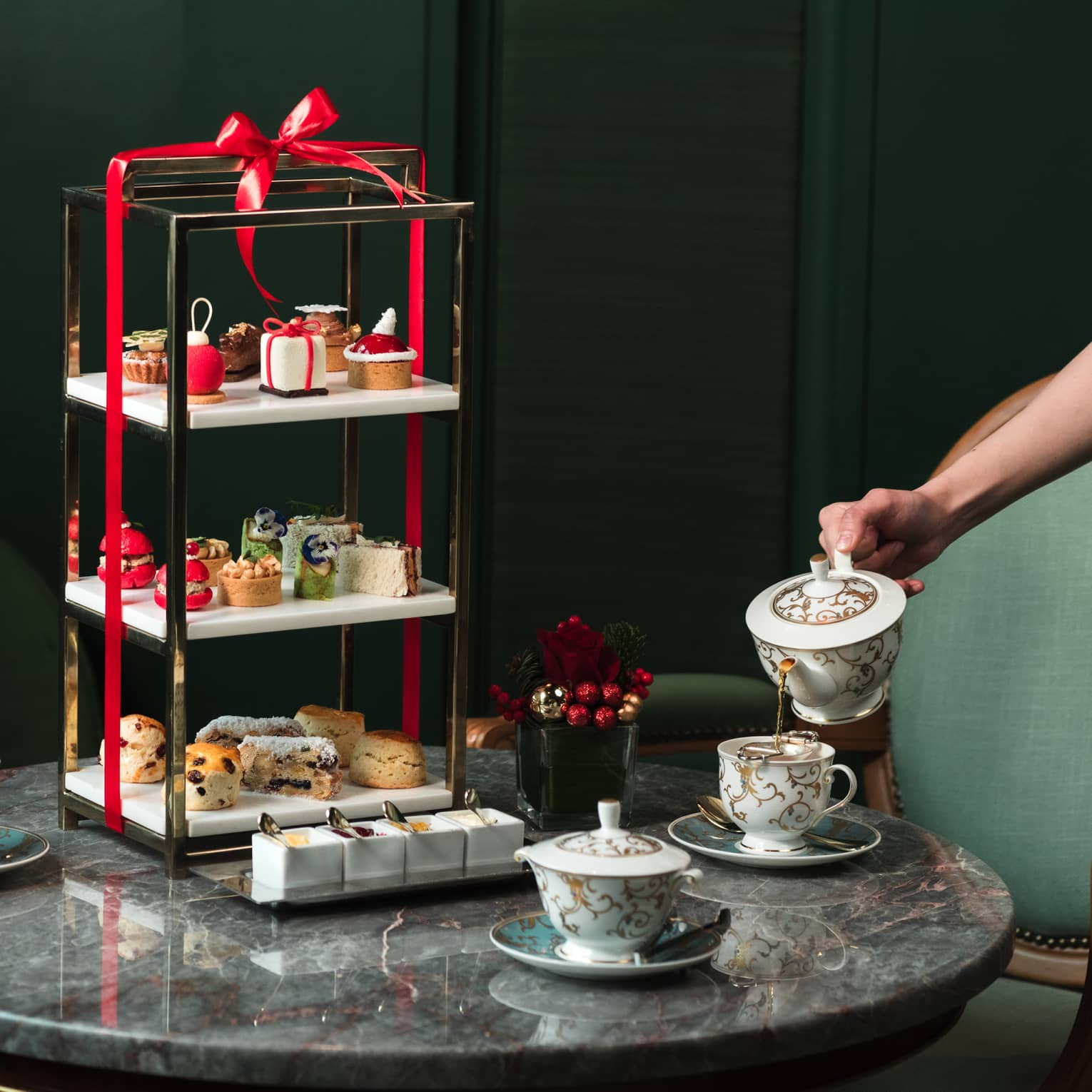 A round grey marble table is set with three tea cups and a three-tiered serving platter filled with pastries and wrapped in a red bow