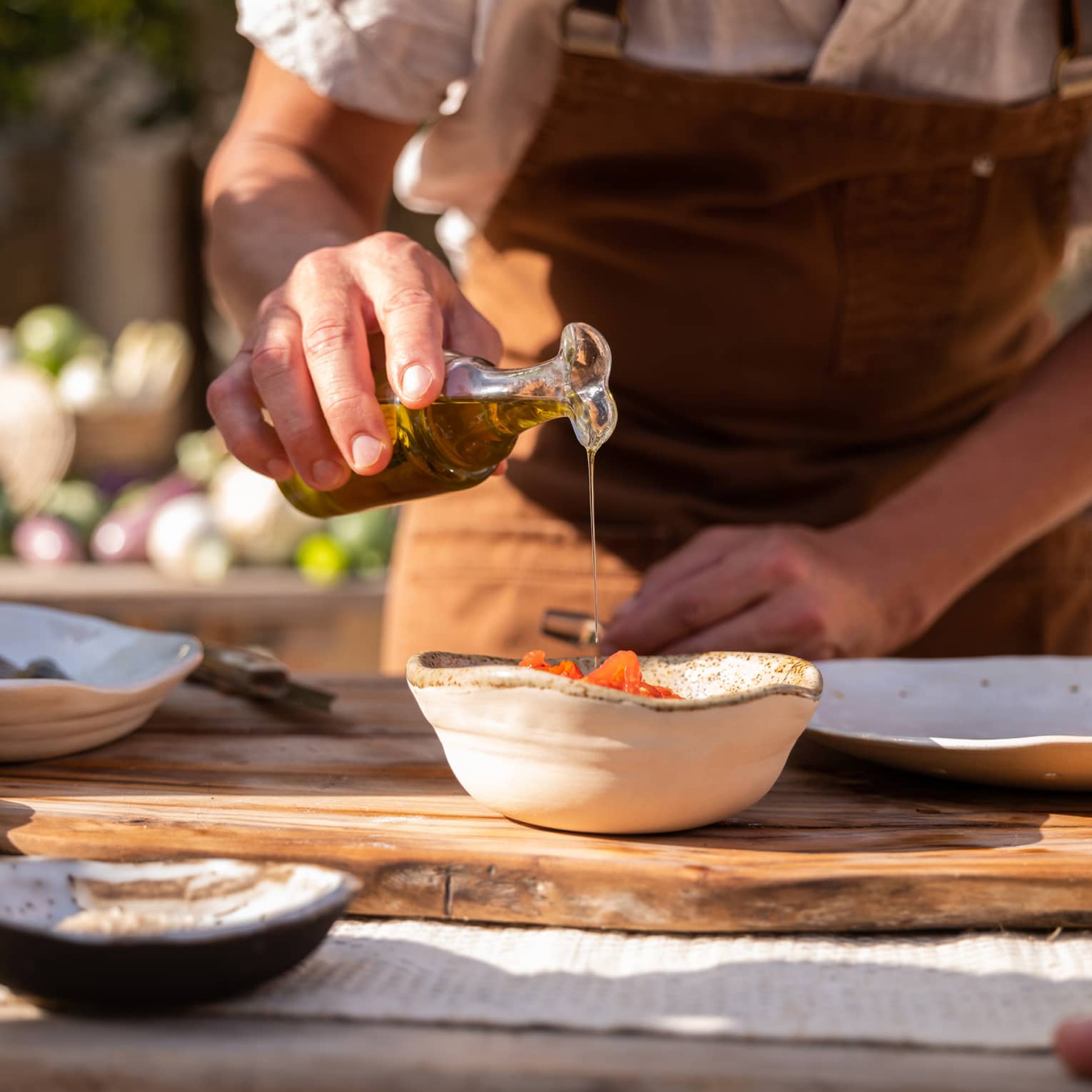 Sunny close-up of a server pouring oil over tomatoes in a bowl set on a wooden cutting board amid dishes and fresh bread.