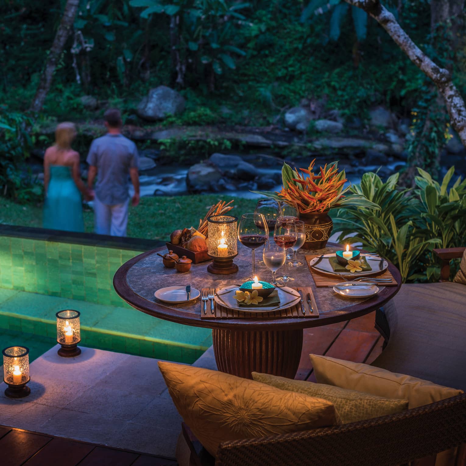 Glowing candles on private patio dining table at night, couple holding hands in background