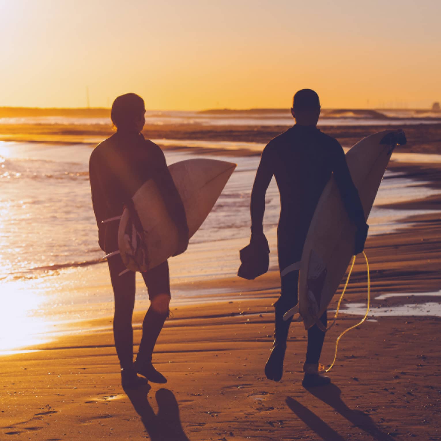 Silhouettes of two people carrying surfboards across beach at sunset