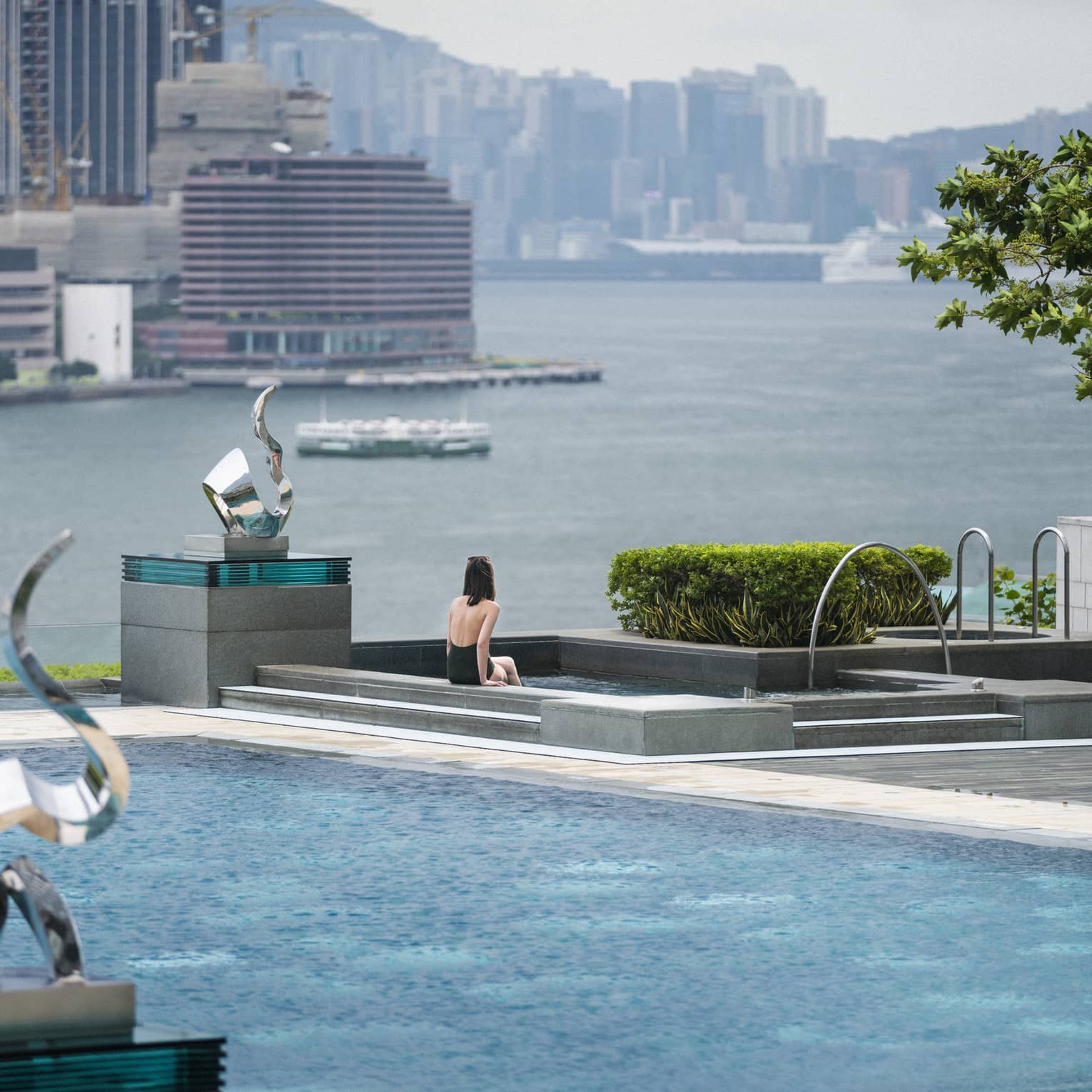 A person is seated at the edge of an outdoor swimming pool, which overlooks a large body of water with city buildings in the background.