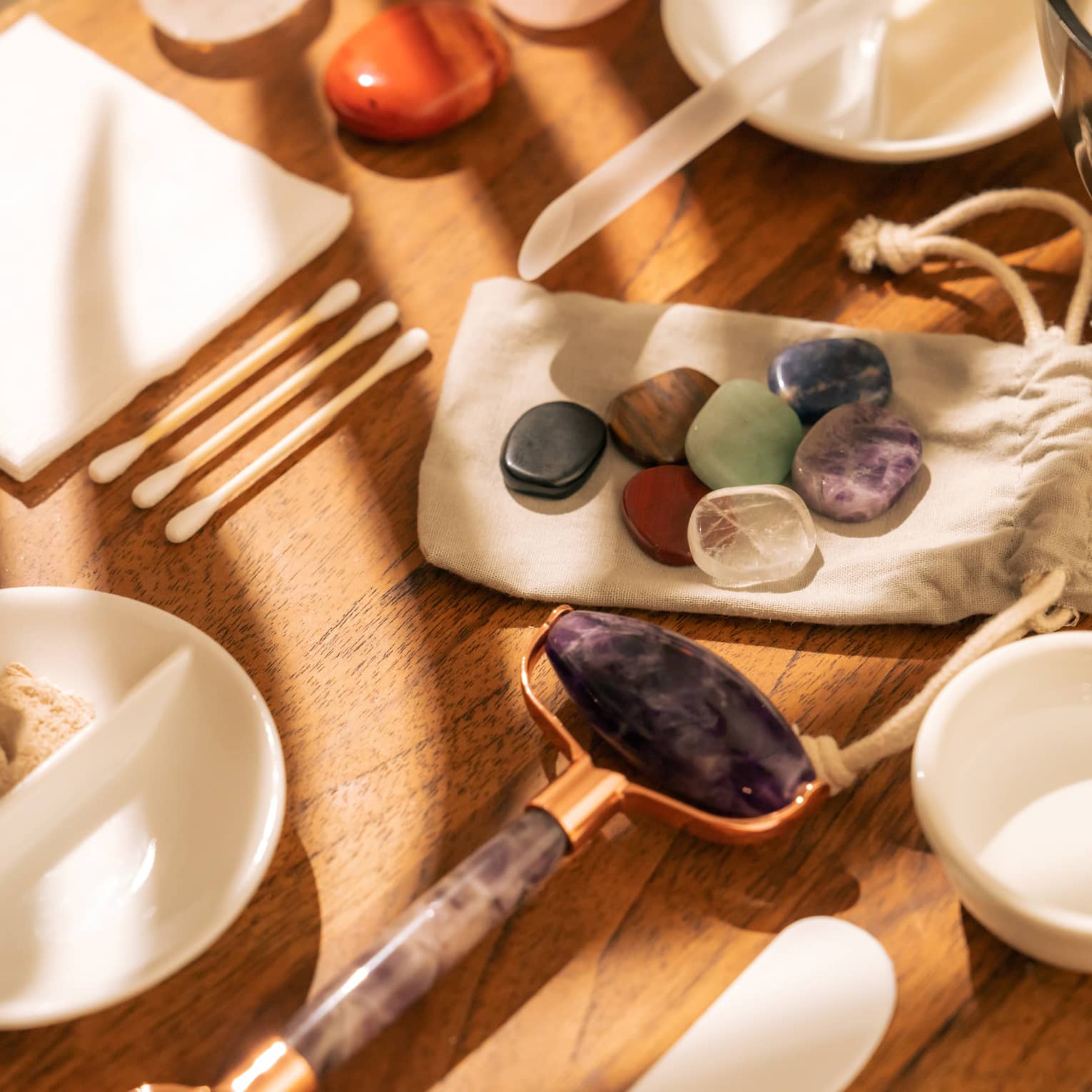 Face roller, gemstones, cotton swabs and a variety of small white bowls and other instruments sit on a sun-lit wooden table