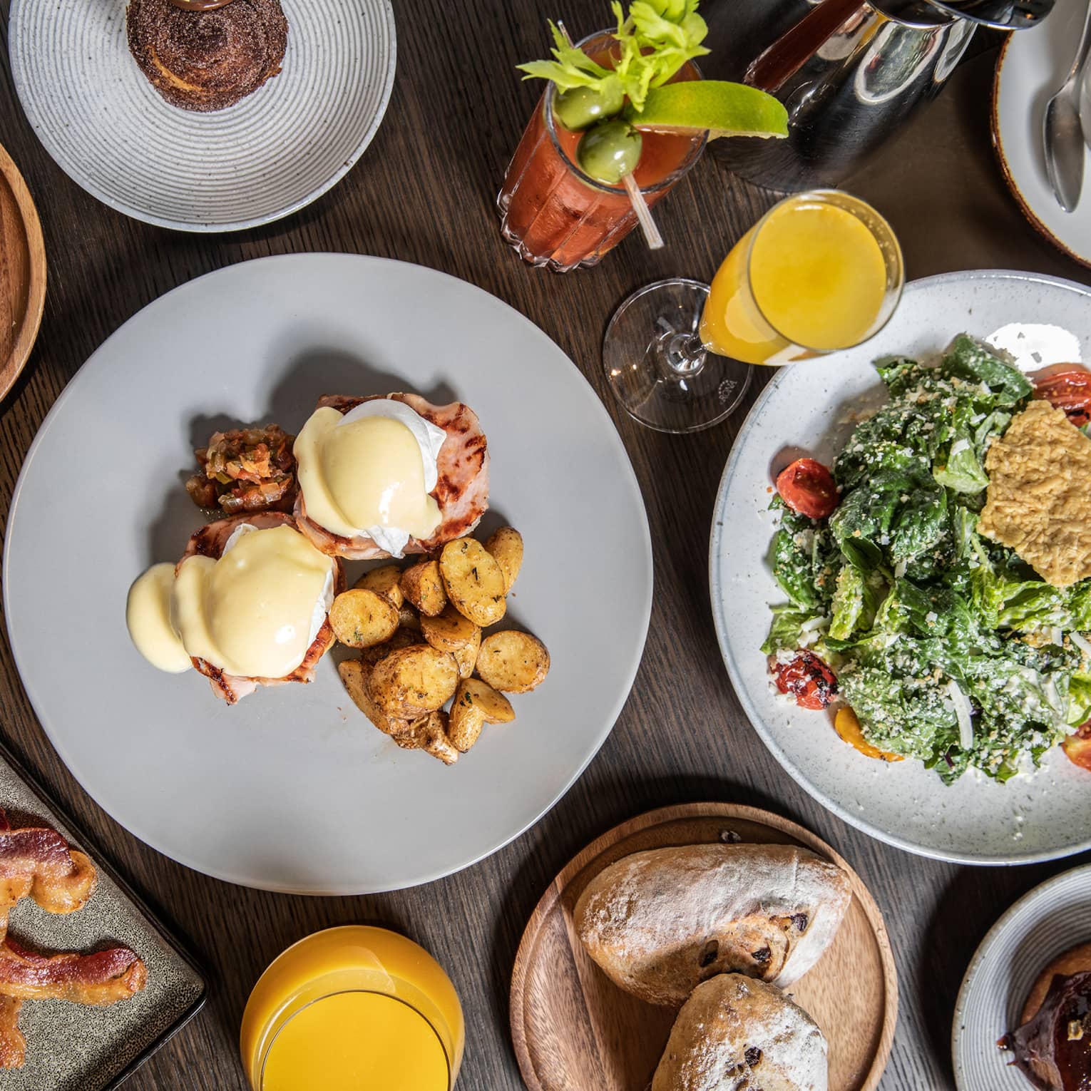 An elaborate brunch, including eggs Benedict, potatoes, bacon, various pastries, salad, a bloody mary and mimosas