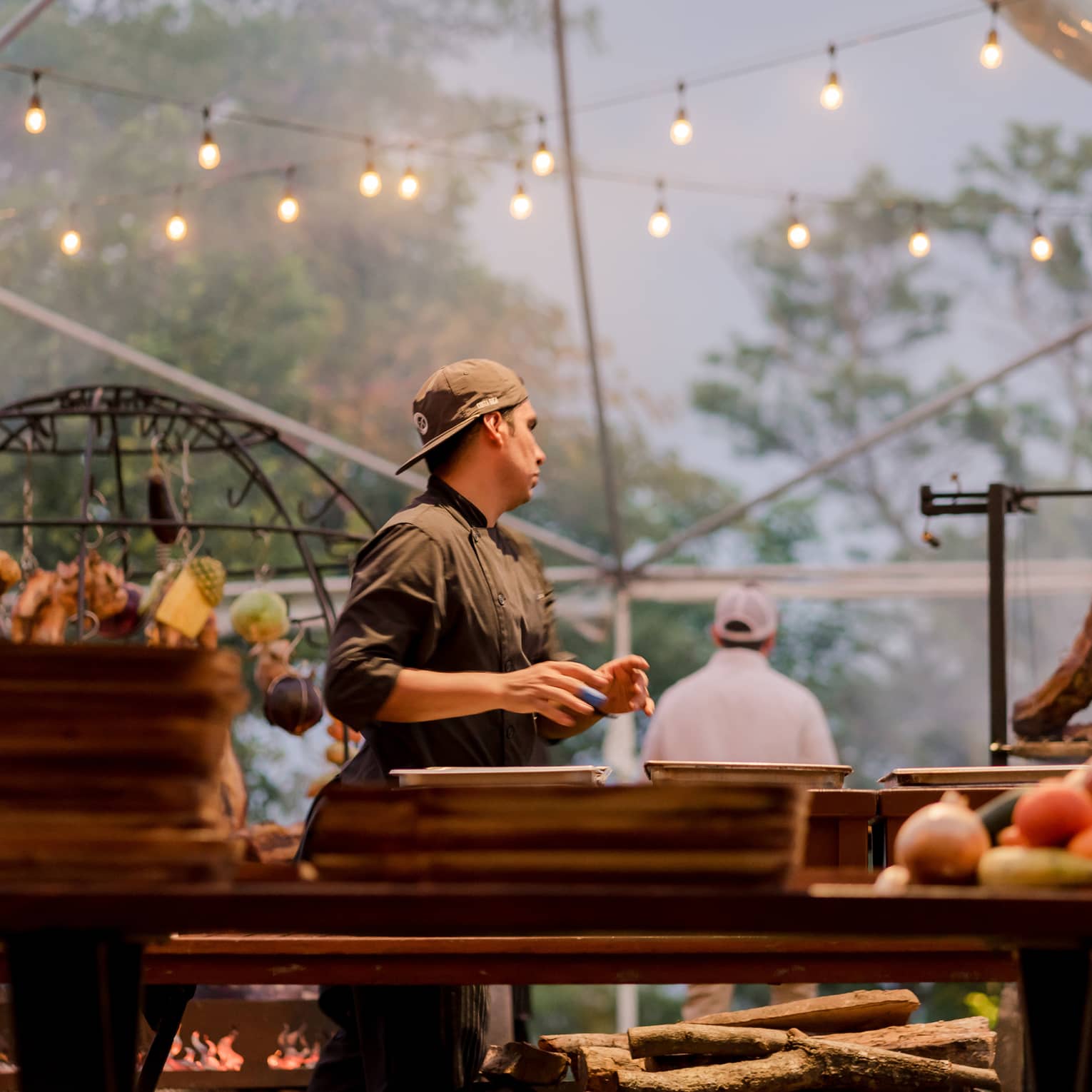Chef wearing a black chef's jacket stands a grilling station beneath two strings of twinkling lights