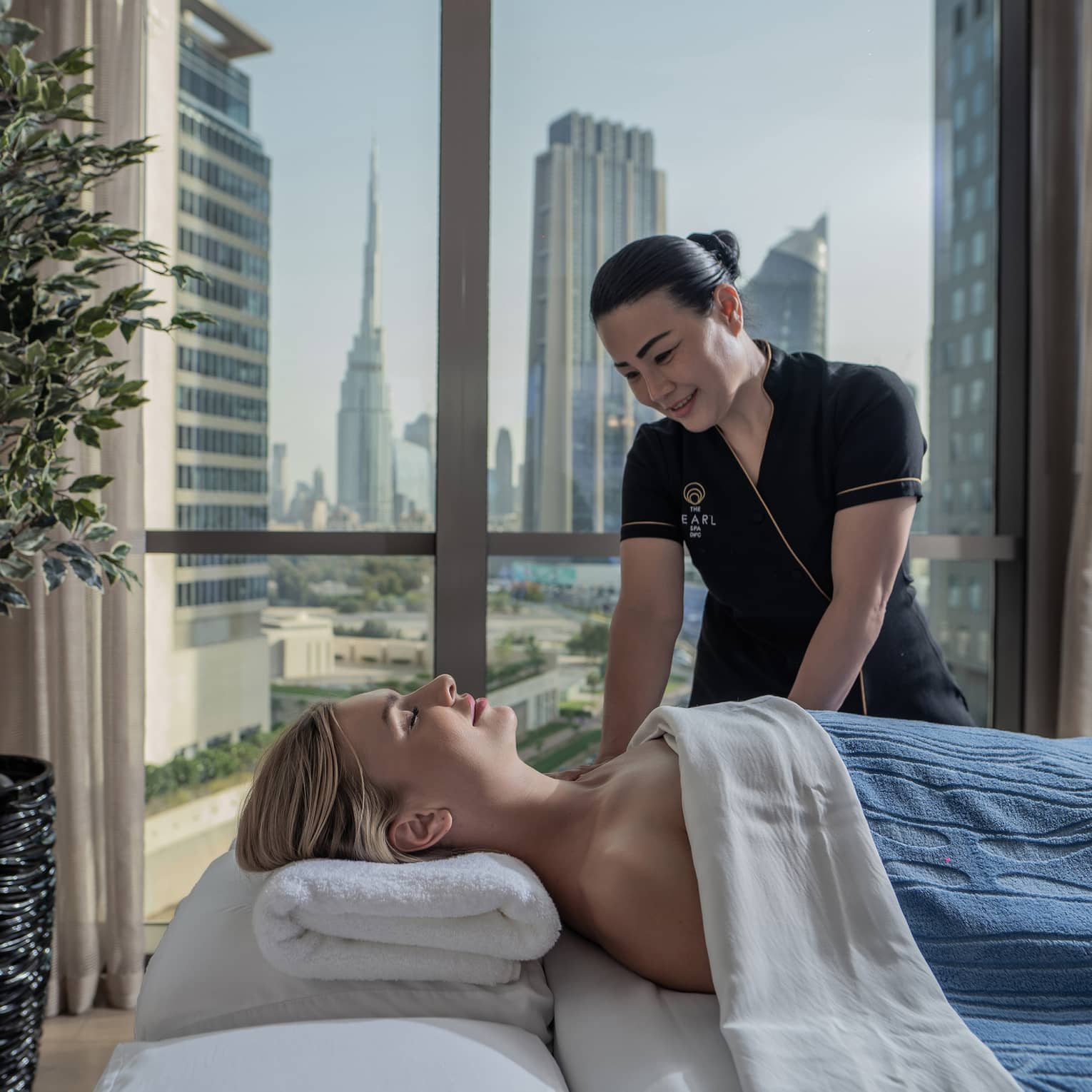 A guest enjoys a massage from a Hotel employee. Behind them, a window frames tall glass skyscrapers and clusters of trees.