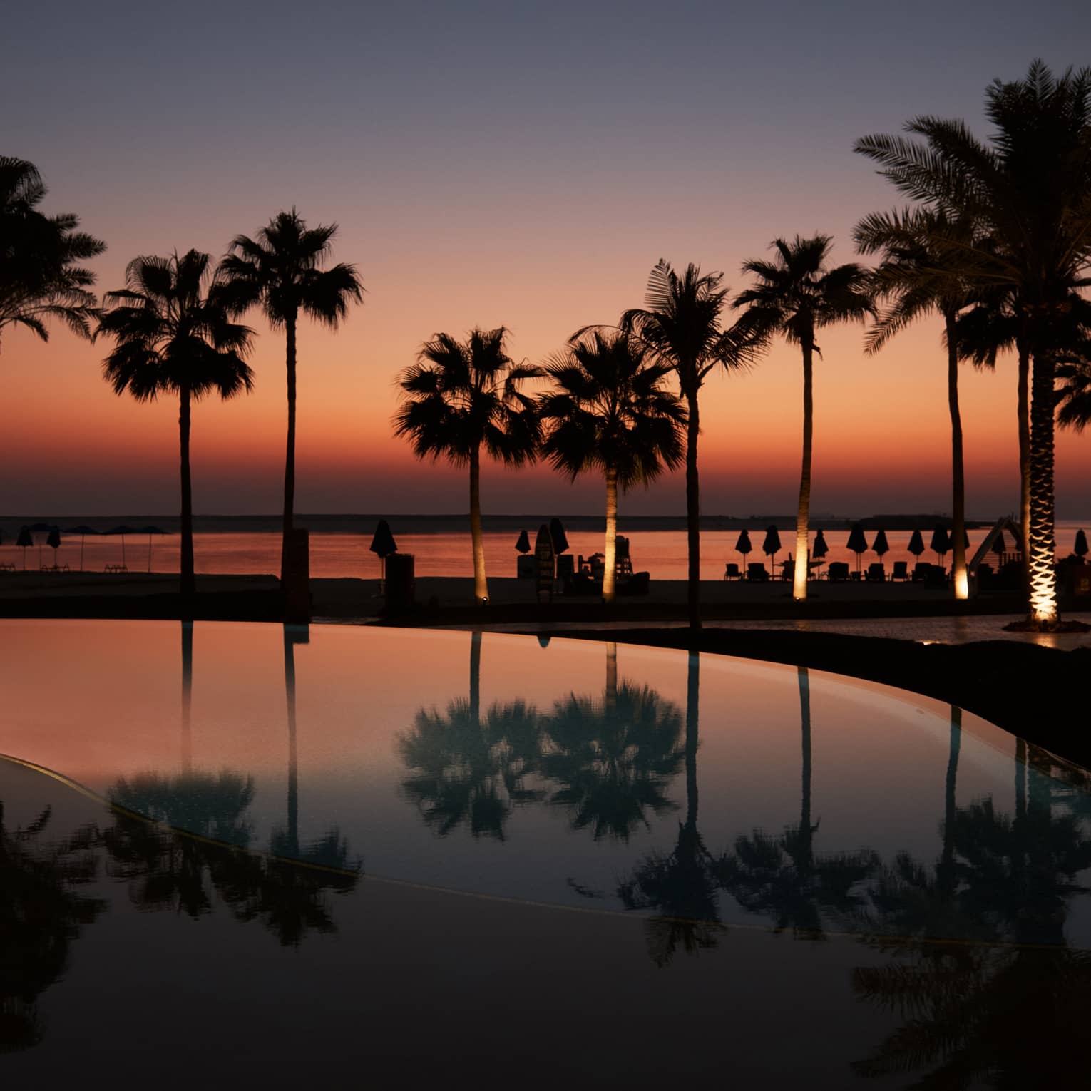 A palm tree?lined pool with the beach in the background all lit under a dark orange and blue sky as the sun sets