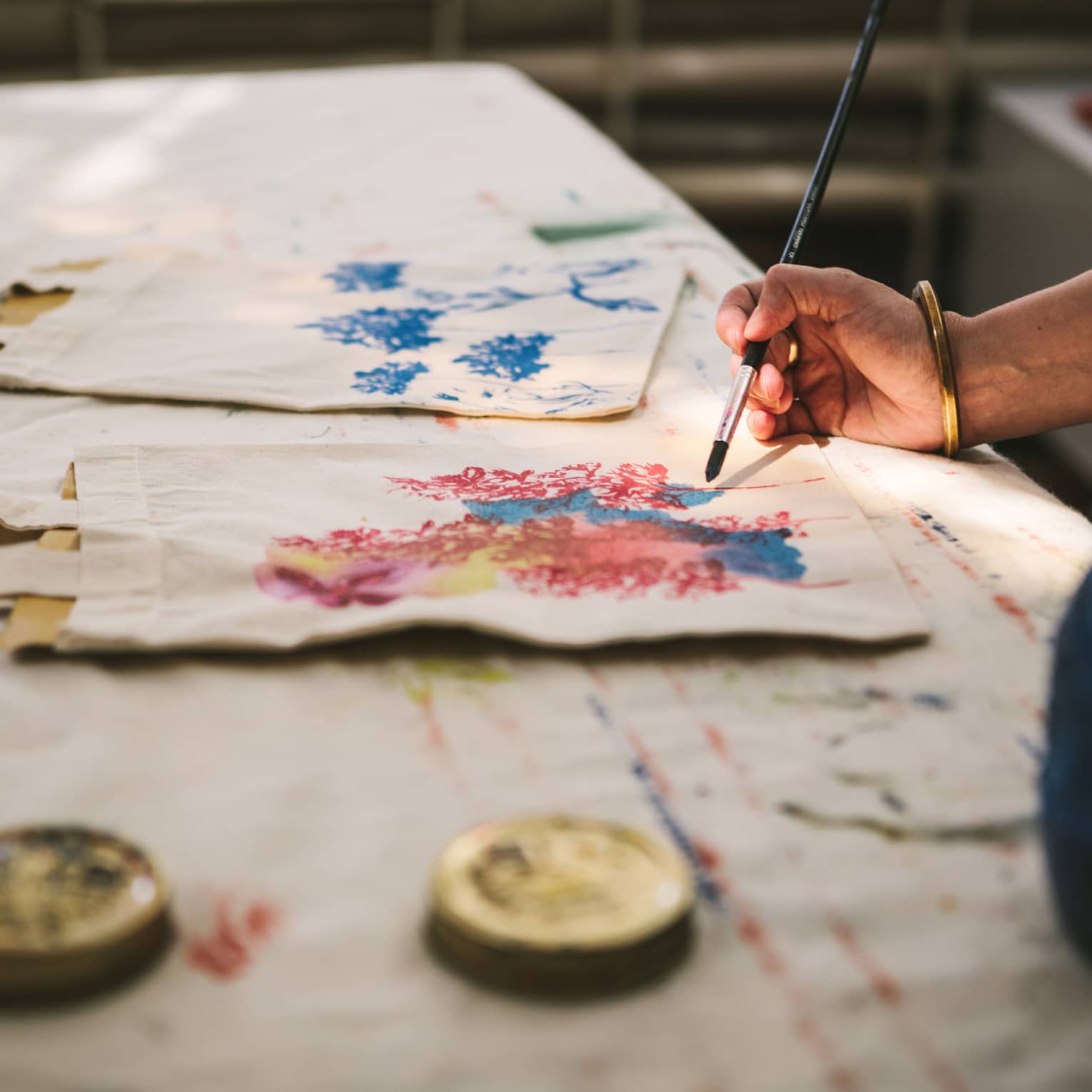 Close-up of a gold-bangled hand wielding a long brush to paint an intricate pattern on a cloth tote bag.