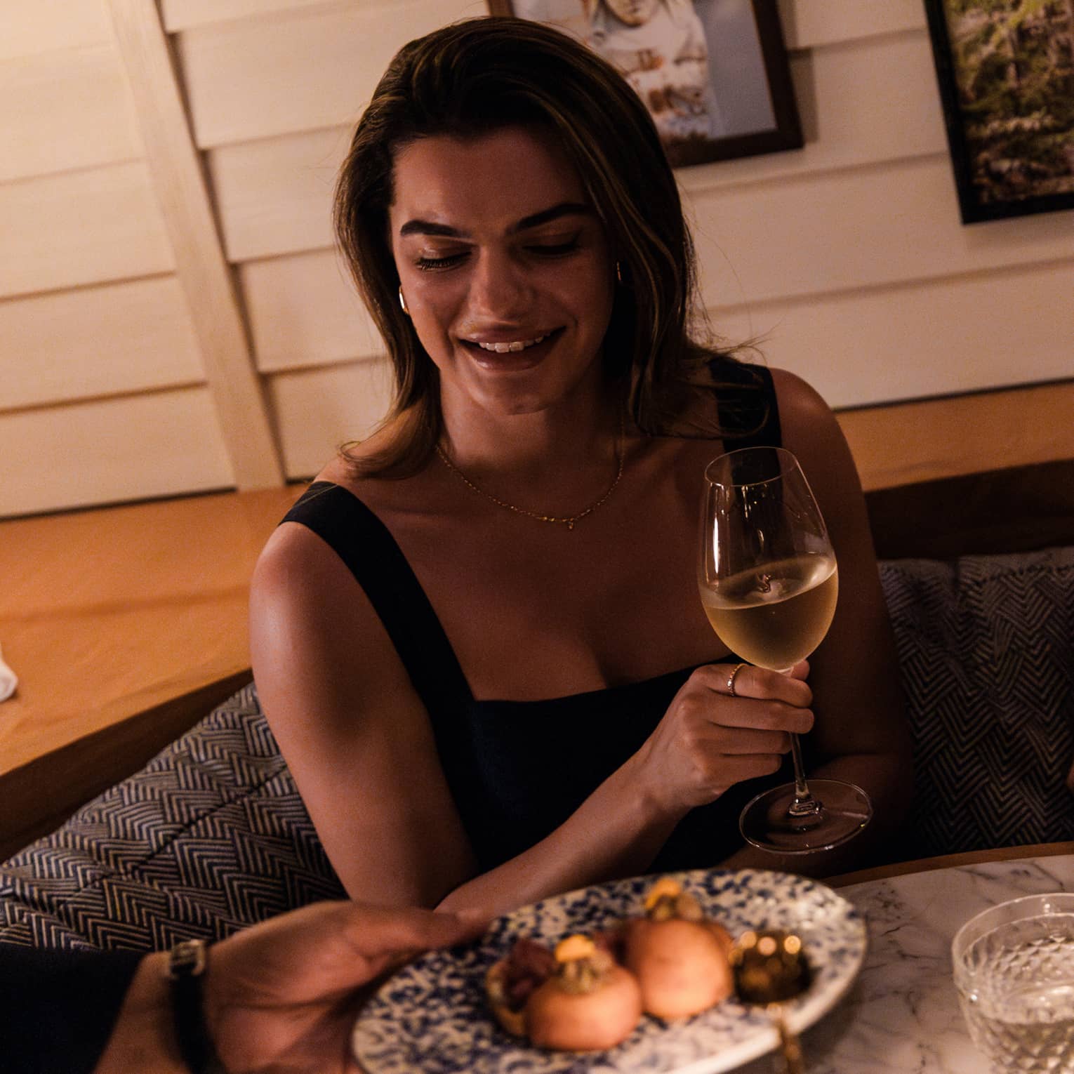 Guests sit at a booth table in a dining space. A person is handing the guests a plate of food, while the guest holds a glass of wine in a stemmed glass.