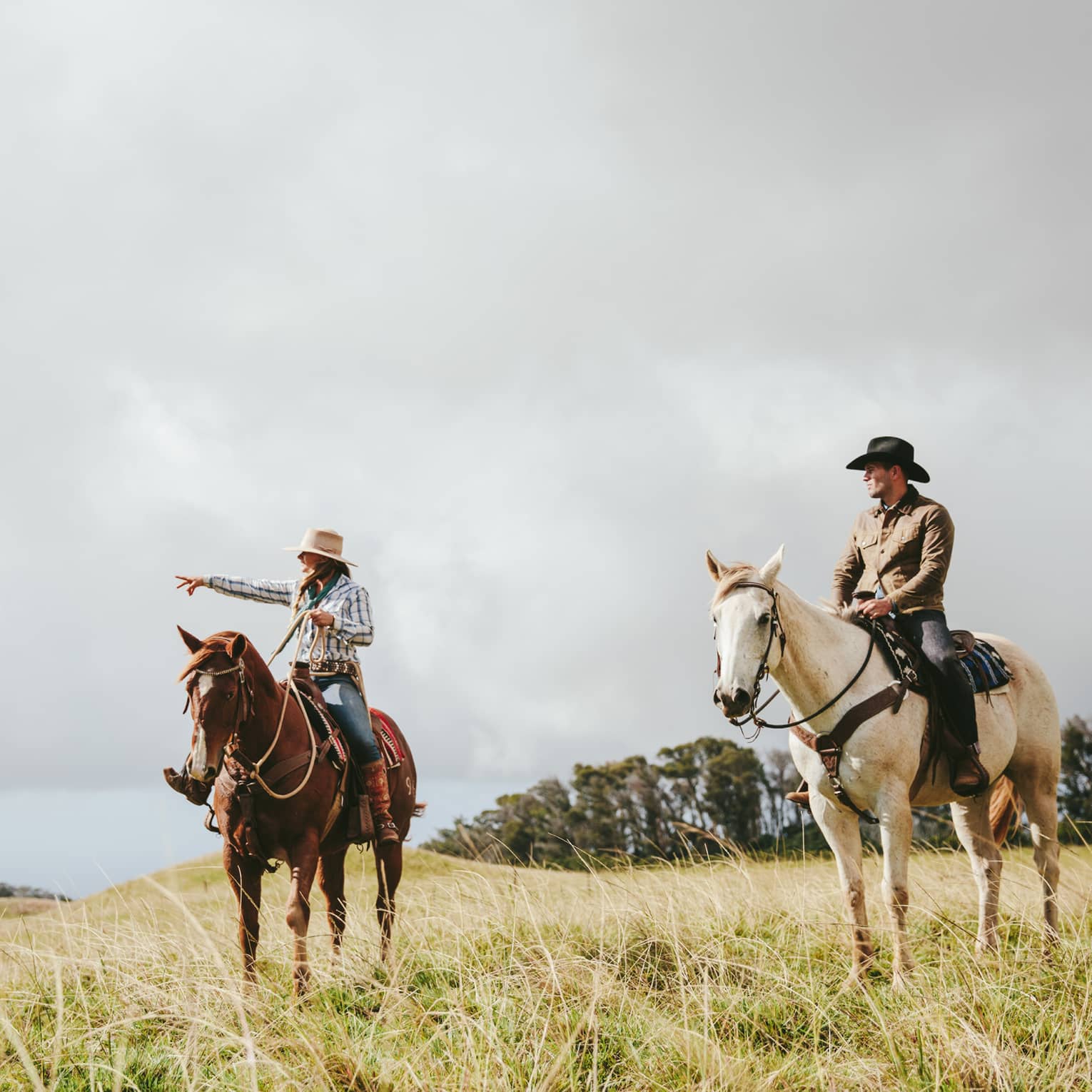 A couple riding horses, looking out at the long grass fields, with grassy hills and trees in the distance on a cloudy day.