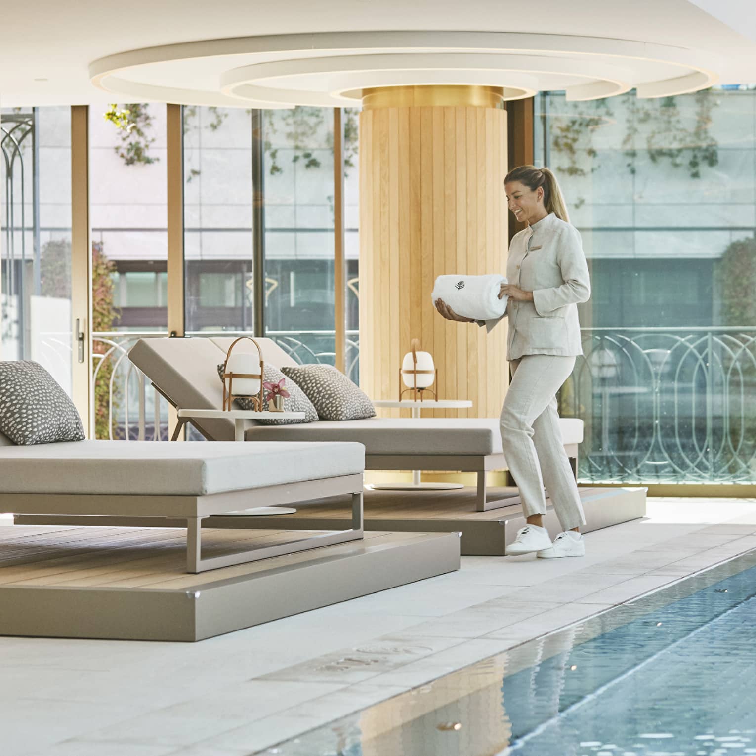 A woman carrying white towels to cushioned lounge seat next to indoor pool