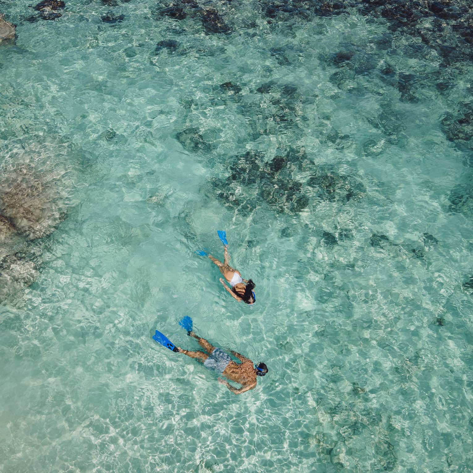 Aerial view of two snorkellers wearing blue flippers in an expanse of shallow, turquoise waters rippling over the reef.