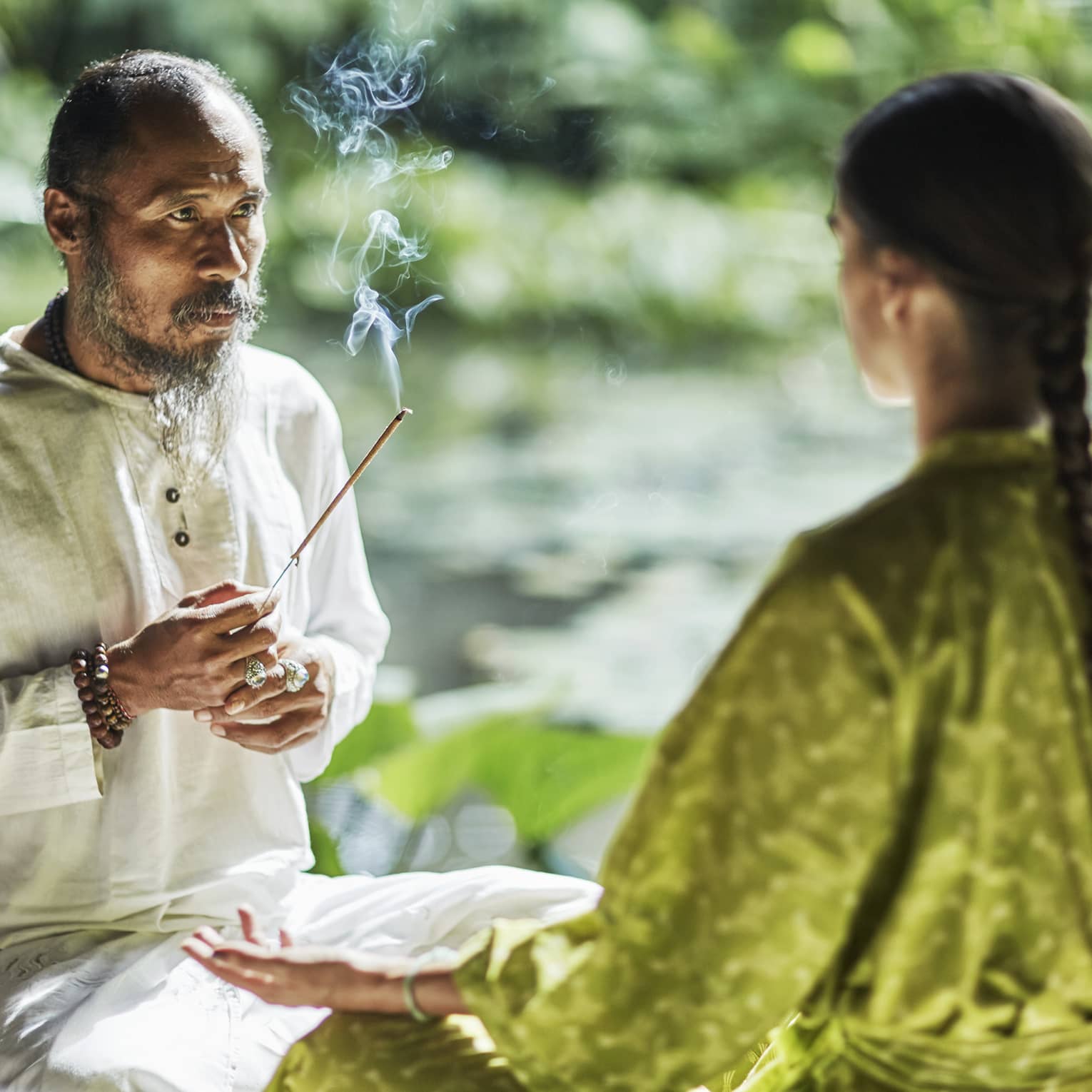 Set against a backdrop of greenery, Djik Dewa holds a stick of burning incense before a meditative cross-legged guest.
