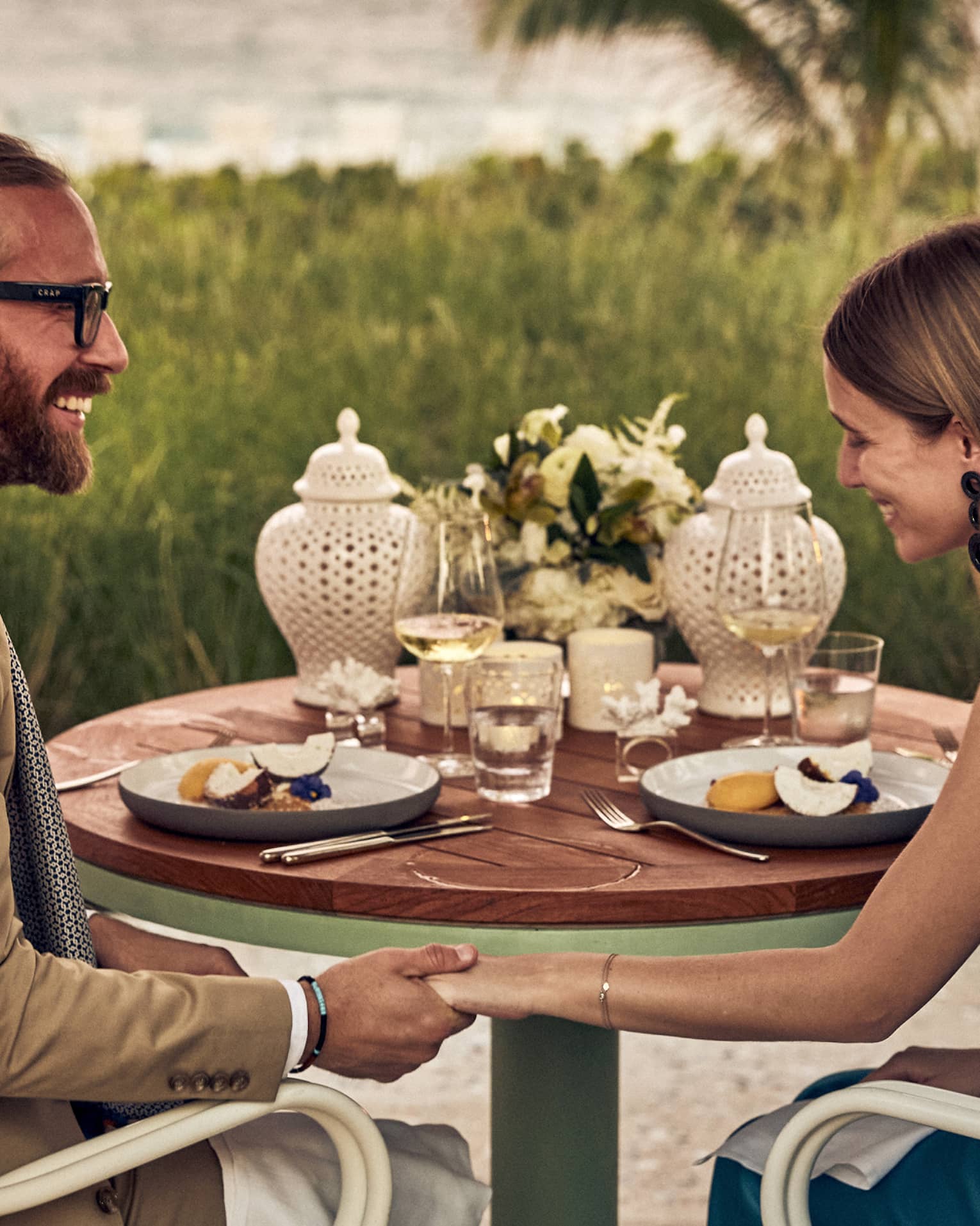 Smiling couple holds hands at dinner set with fruit plates, wine, flowers and two white perforated porcelain candle holders.