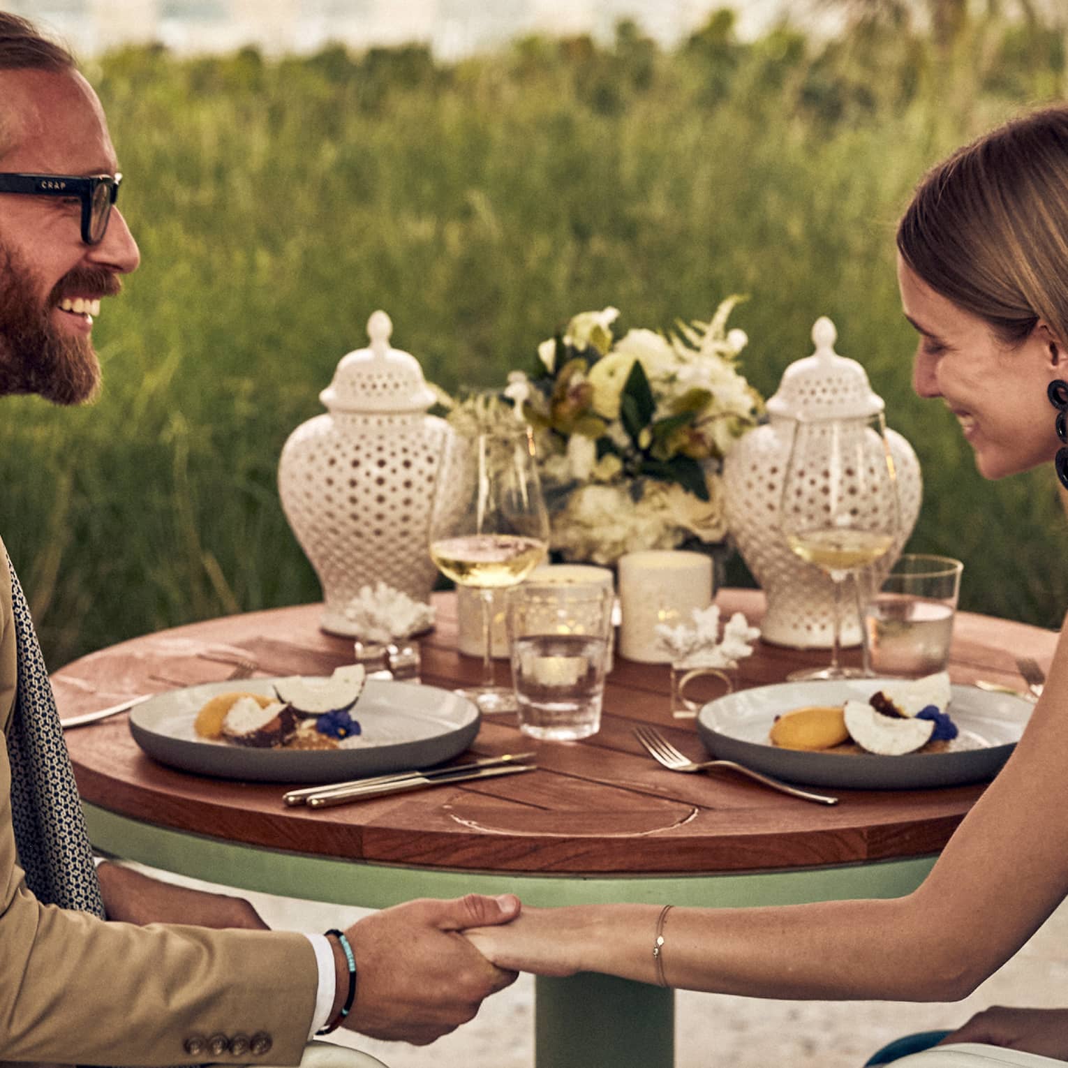 Smiling couple holds hands at dinner set with fruit plates, wine, flowers and two white perforated porcelain candle holders.