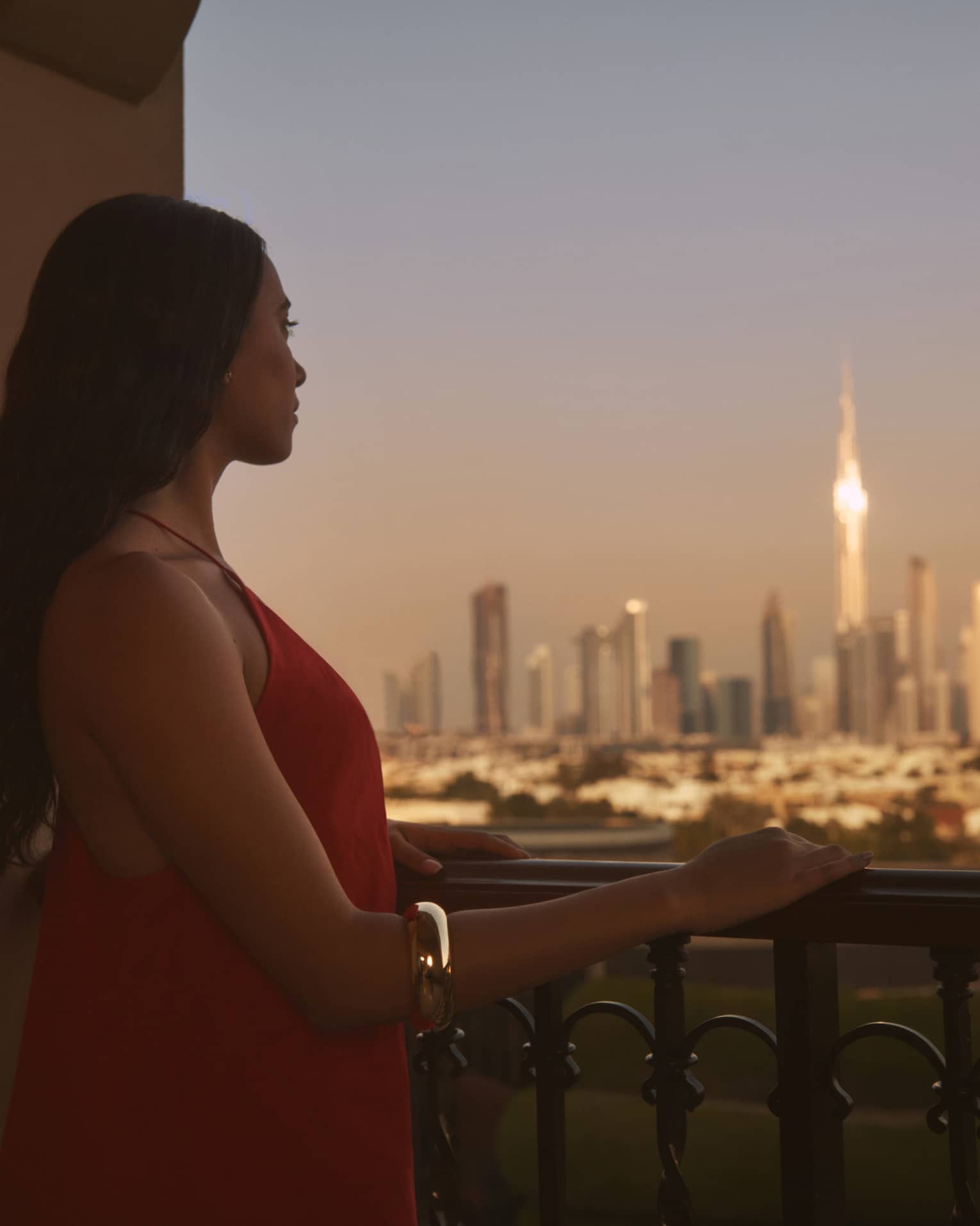 Person with long brown hair standing on balcony with hand resting on railing, looking out to a city at dusk