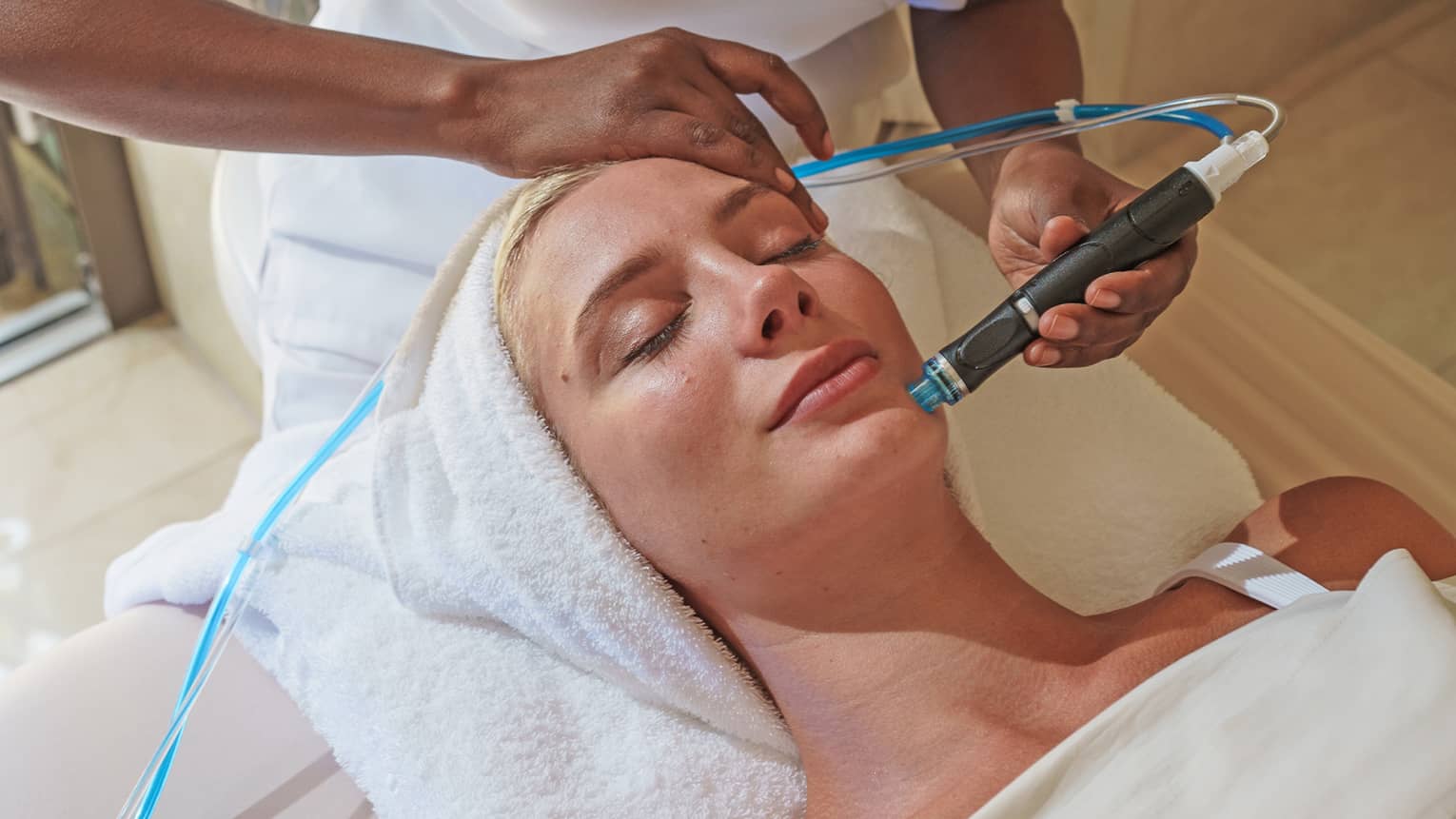 Spa staff holds a blue and black facial tool over woman's face as she lays on massage bed 