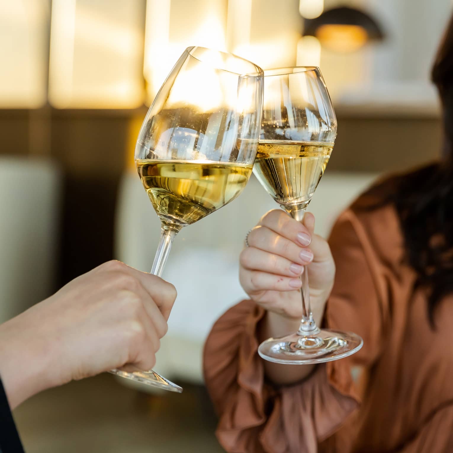 Close-up of two hands clinking glasses of white wine together; beyond, a person in flowing blouse and long brown hair smiles.
