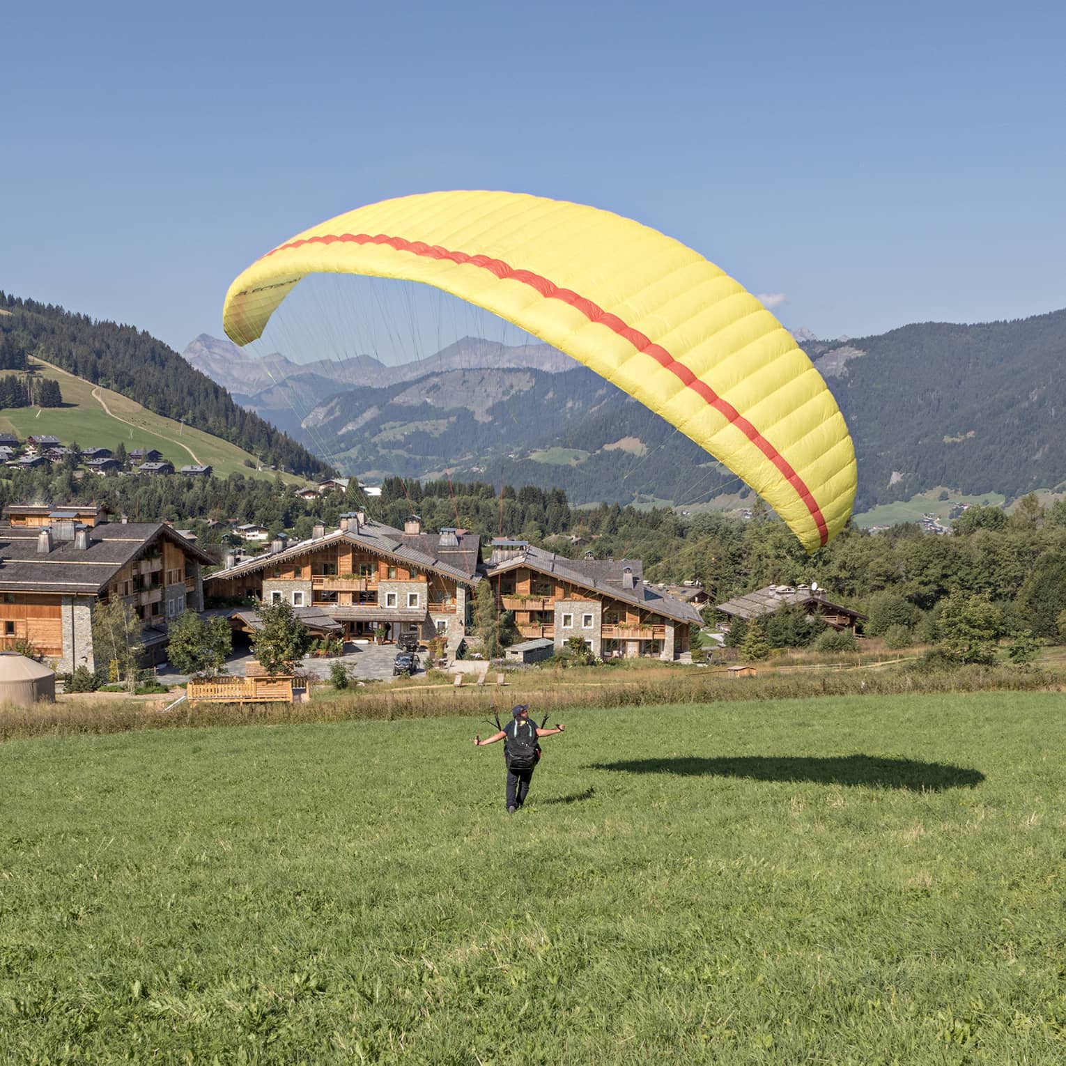 Person on meadow holding yellow parachute in air, facing rustic Megève hotel and verdant mountains