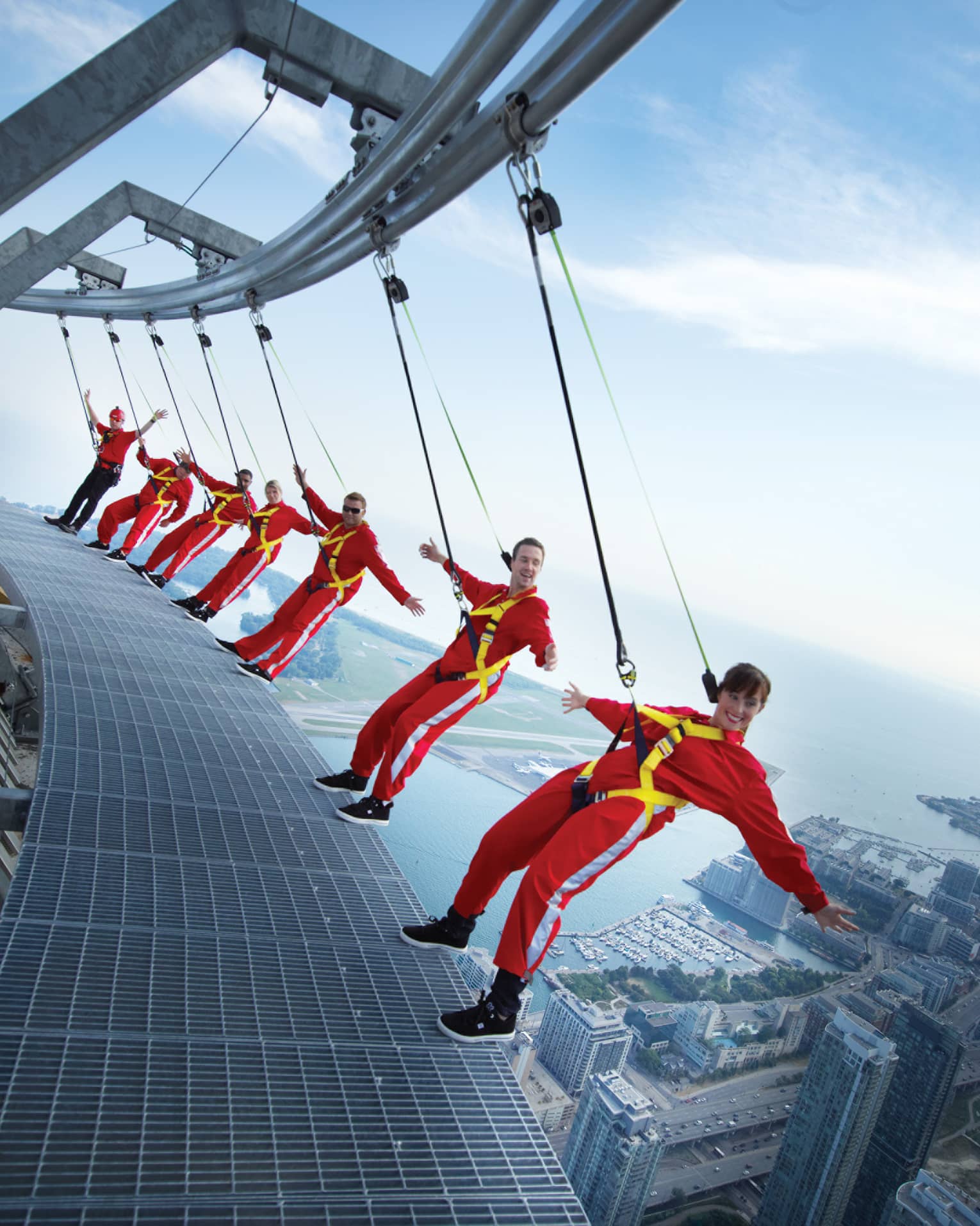 People wearing red jumpsuits, harnesses lean back over edge of CN Tower high above city