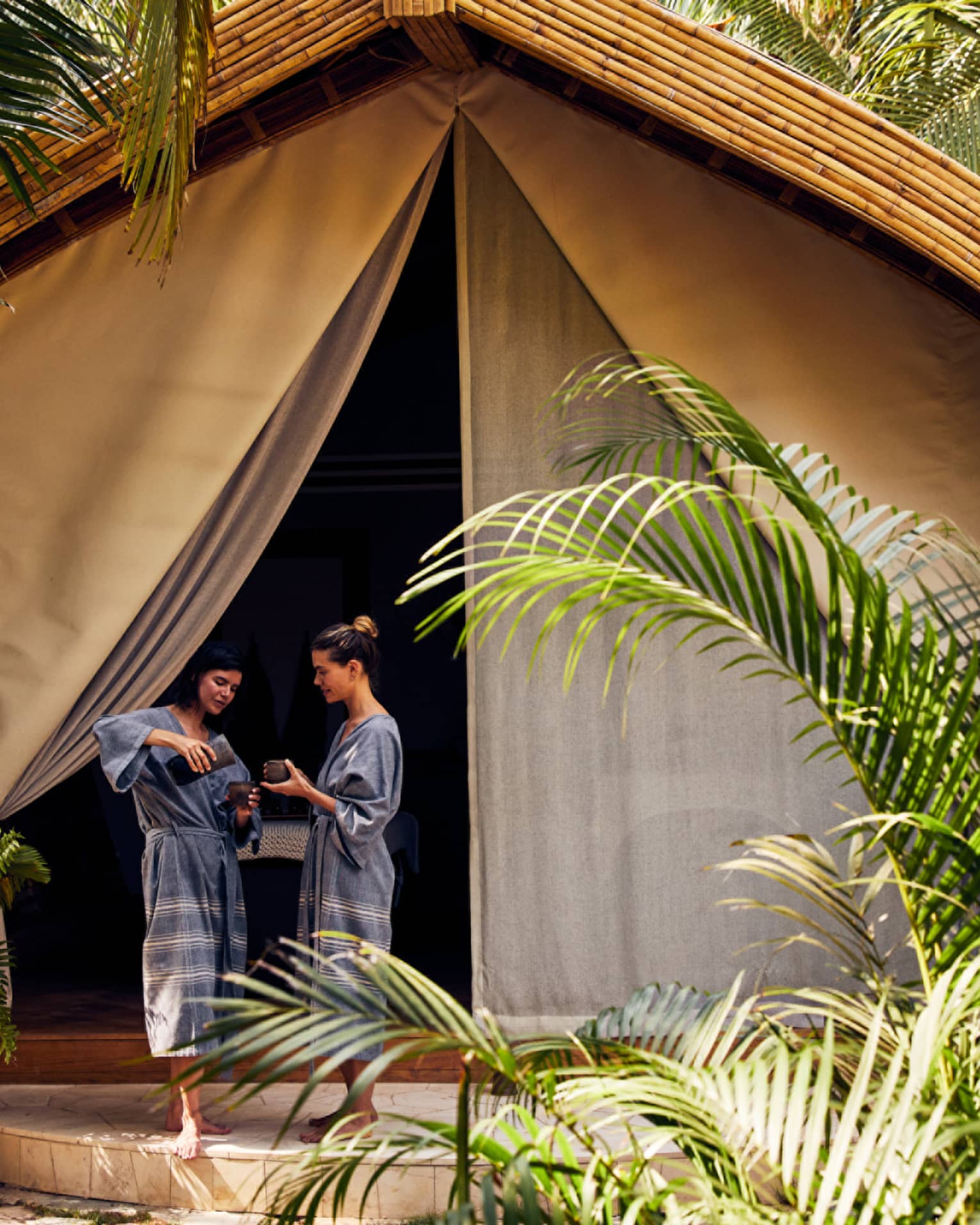 Two women in spa robes entering a spa tent surrounded by palm trees.