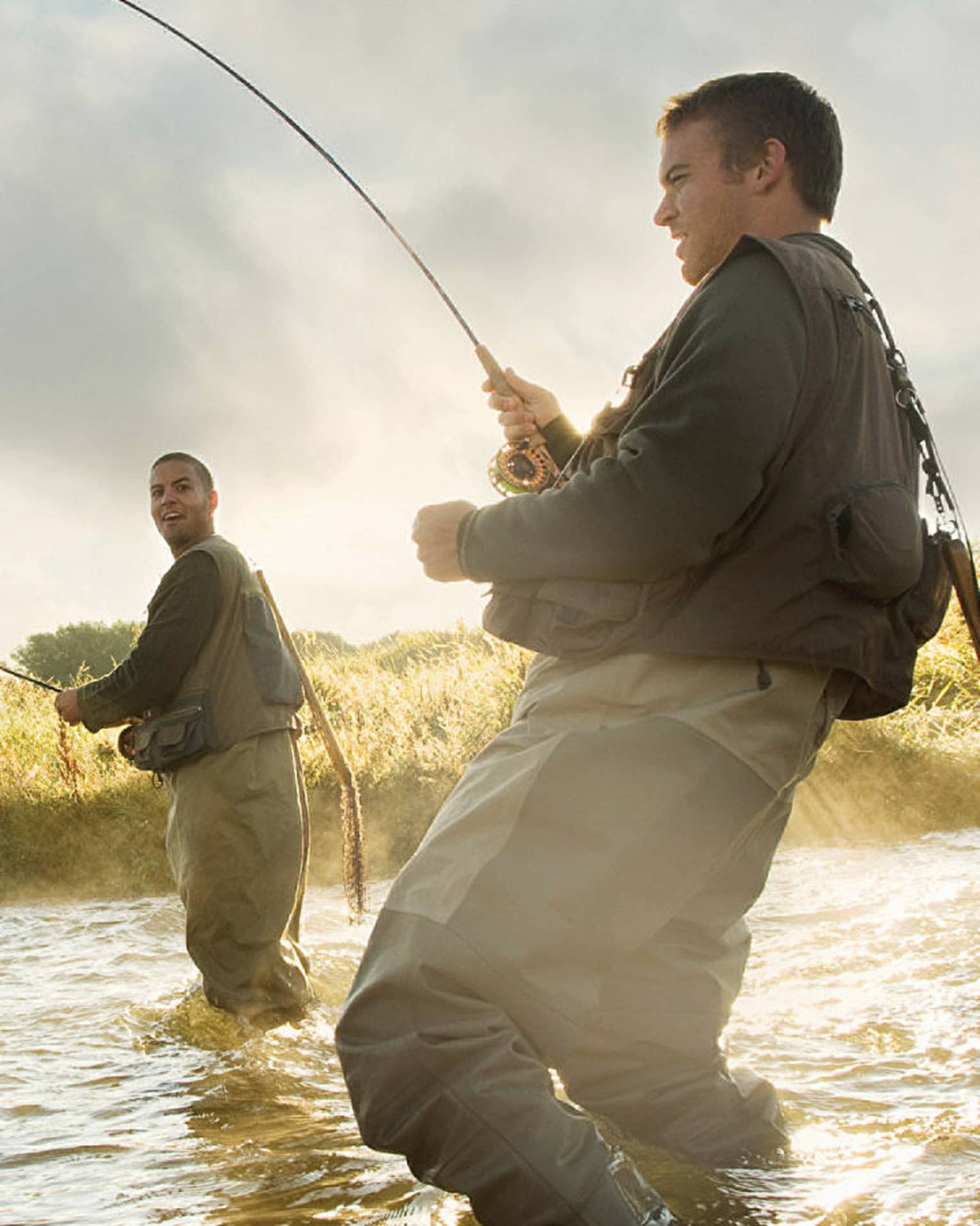 Two people fly fishing in a river, standing knee-deep in the water, surrounded by mist and greenery.