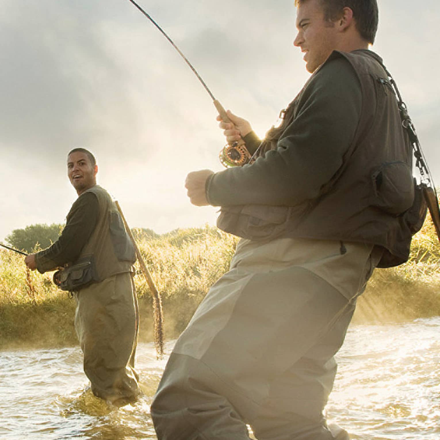 Two people fly fishing in a river, standing knee-deep in the water, surrounded by mist and greenery.
