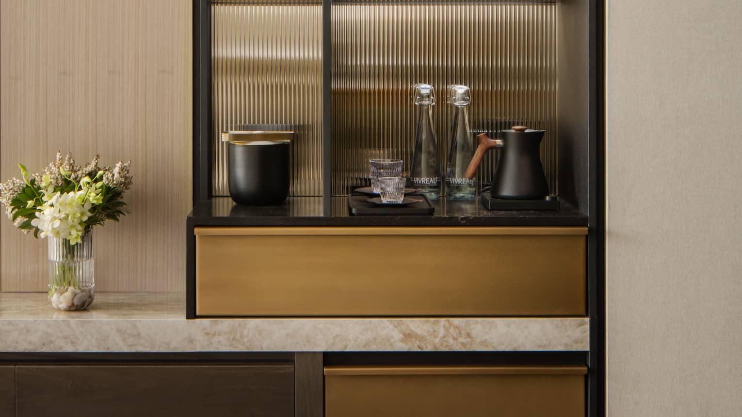 Stylish minibar with glass shelves displaying wine, glasses, and decorative items, next to a TV and vase with fresh flowers