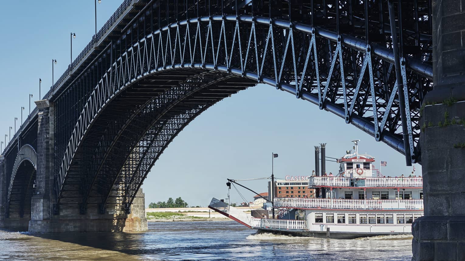 A river boat going under a large metal bridge on a bright day on a river.