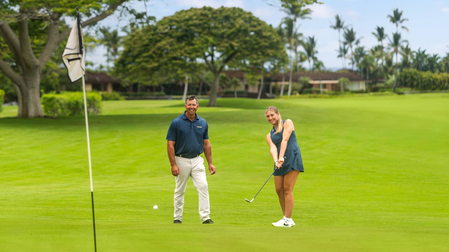 Two people on a golf course with one practicing her short game. 