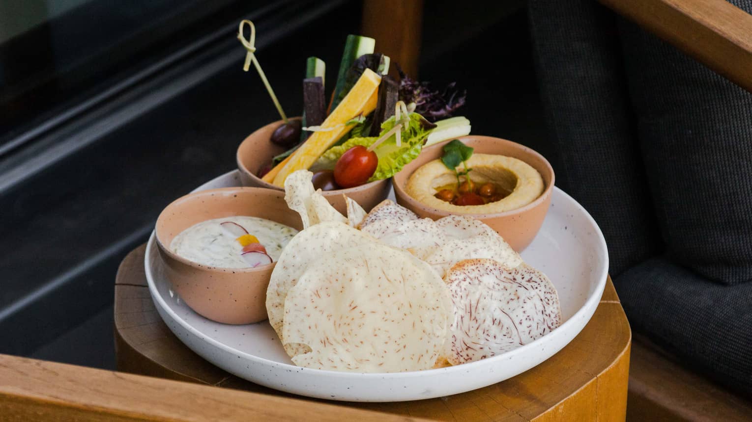 A white round platter is filled with three small clay bowls filled with hummus, dip and an assorment of vegetables with round chips on the side