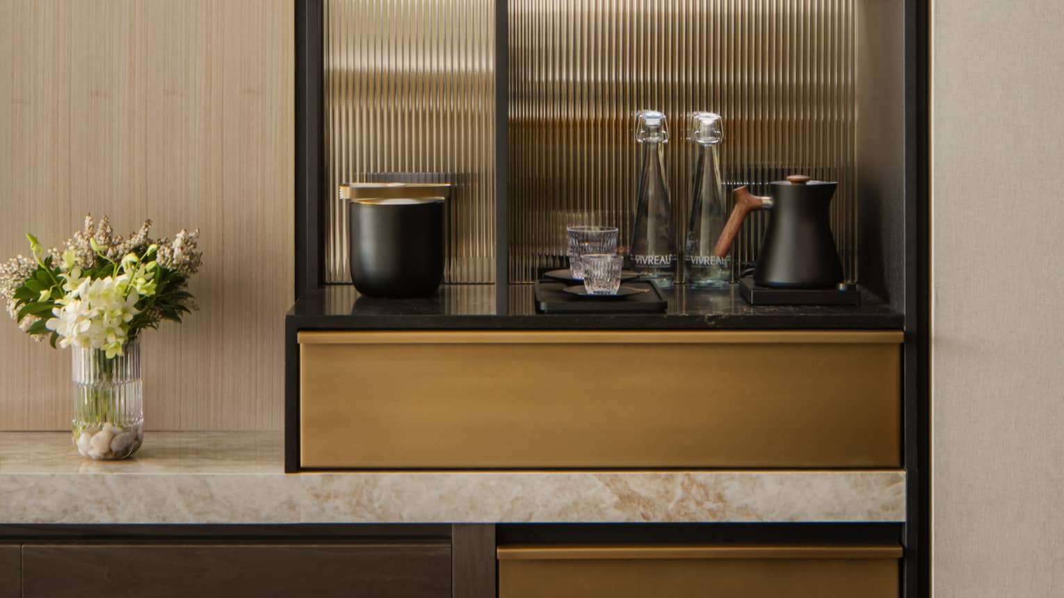 Stylish minibar with glass shelves displaying wine, glasses, and decorative items, next to a TV and vase with fresh flowers