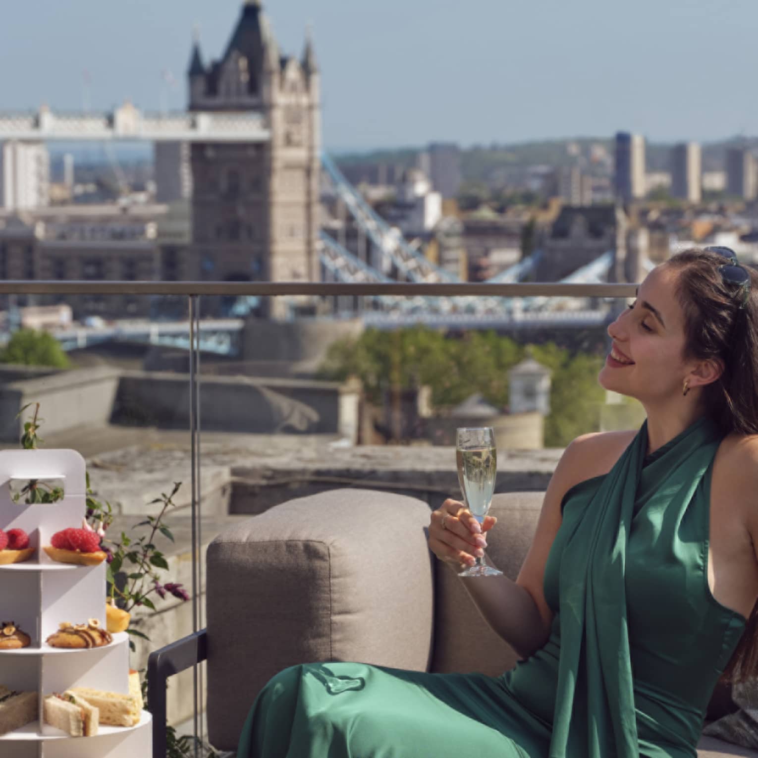 Woman in green dress holding champagne beside tea stand with confections and Tower Bridge in backdrop