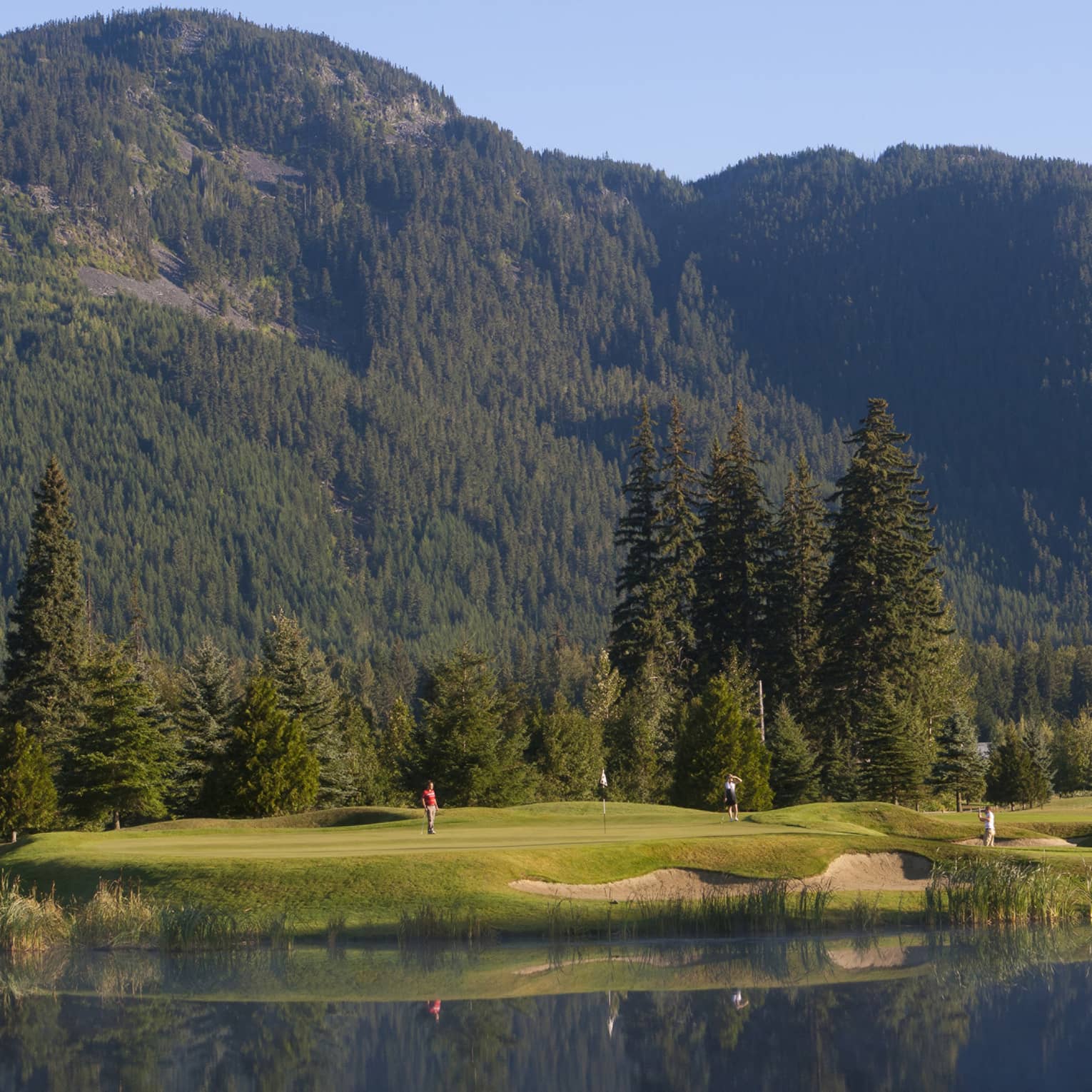 Distant golfers on a green, a pond in front, evergreens behind, dwarfed by a treed mountain filling most of the frame beyond.