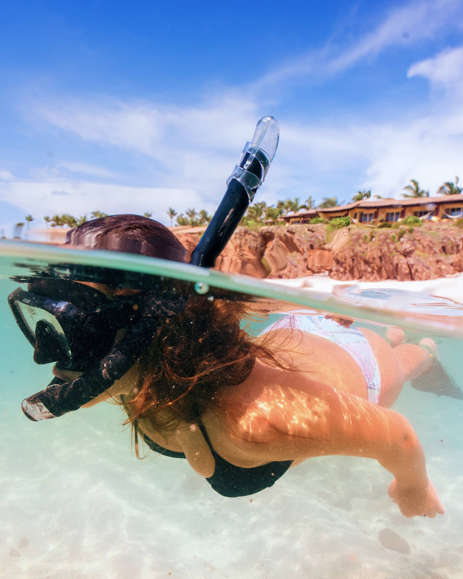 A snorkeller beneath the surface of clear aqua water. Behind, a white sandy beach, swaying palm trees and low-rise buildings.