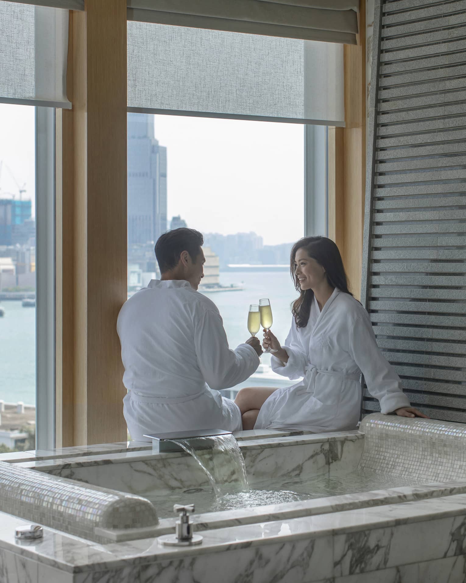 Man and woman clinking champagne glasses next to a private hot tub overlooking city harbour