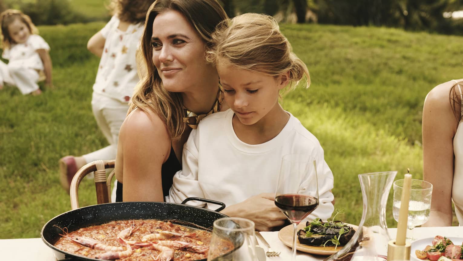 A smiling guest and a child sit together at an outdoor dining table set with a pan of paella, fresh seafood dishes and wine.
