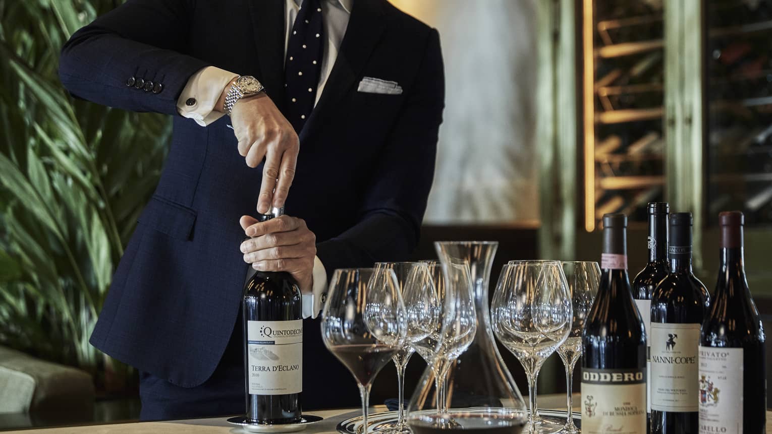 A person opening a bottle of wine, with wine glasses and additional wine bottles beside it
