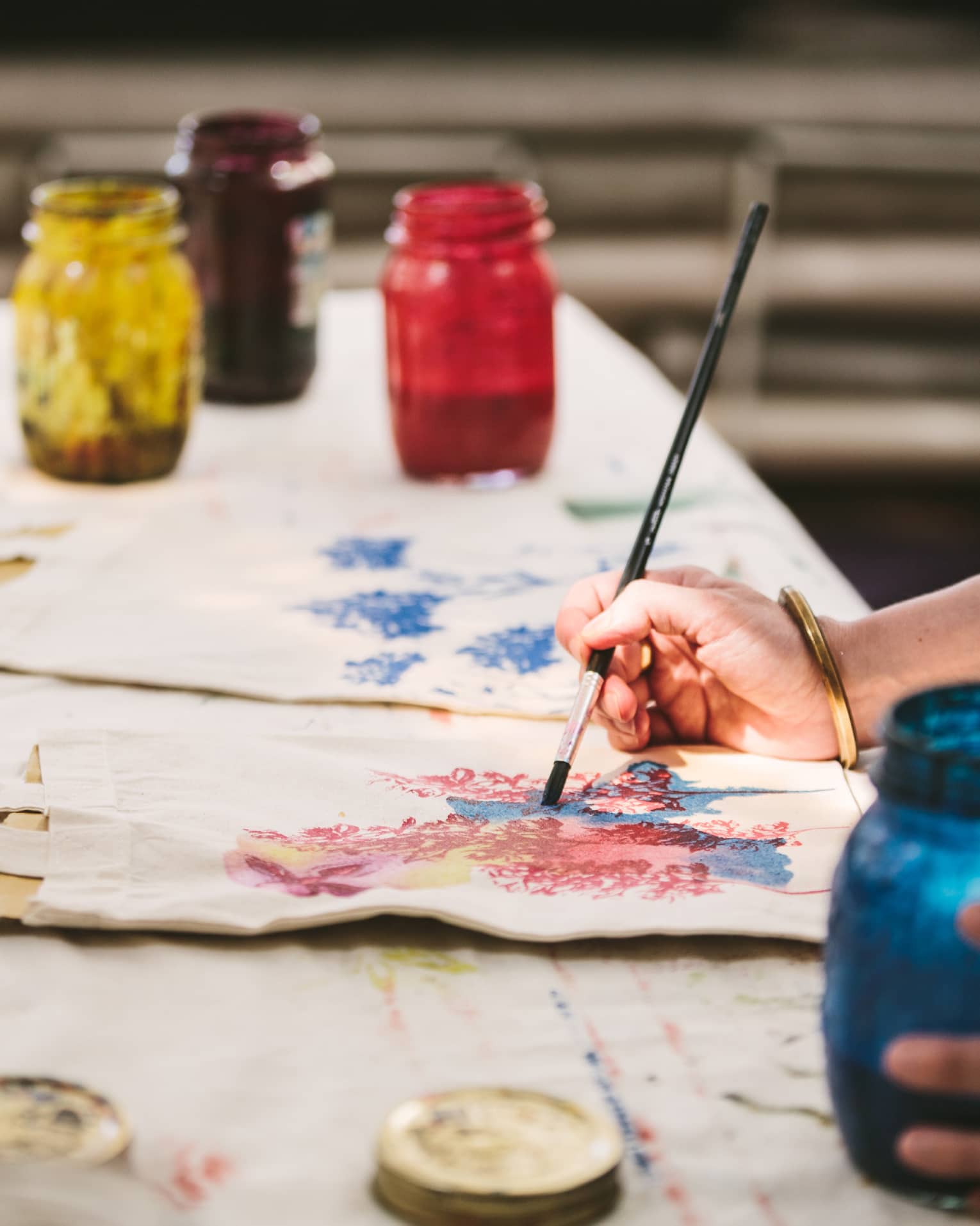 An artist using mason jars filled with red, blue, yellow and black paint to paint images of flowers on canvas tote bags.