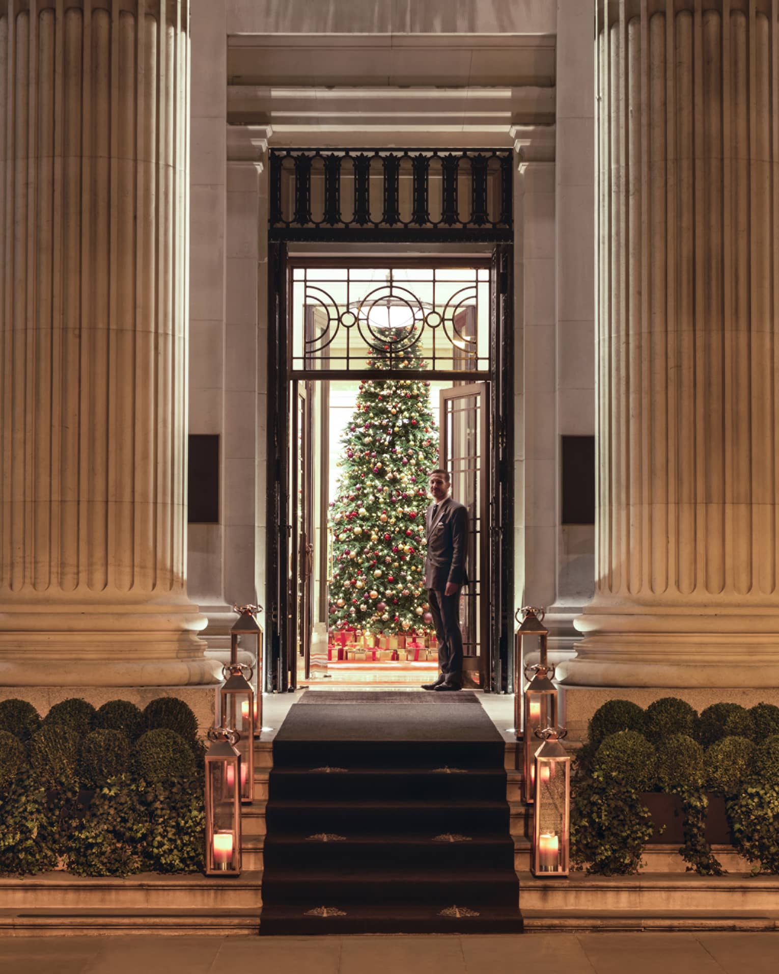 A man waits in a doorway with a Christmas tree behind him. He is centered between two pillars. 