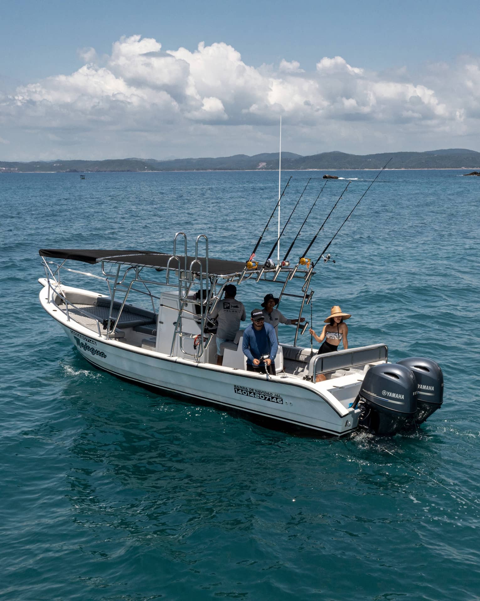 Guest reels in a large yellow fish from the back of a boat in open water near rocky islands, while two friends look on.