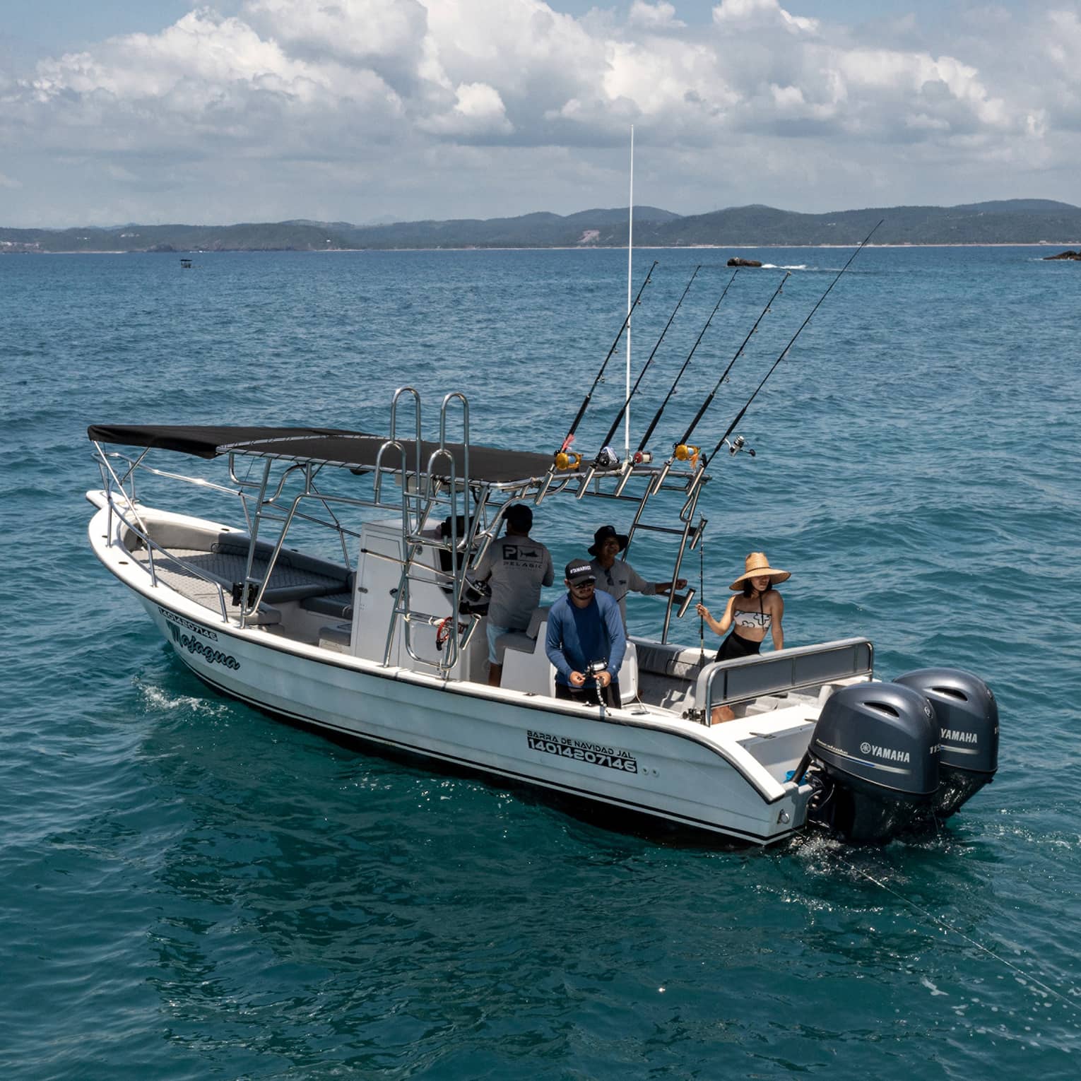 Guest reels in a large yellow fish from the back of a boat in open water near rocky islands, while two friends look on.